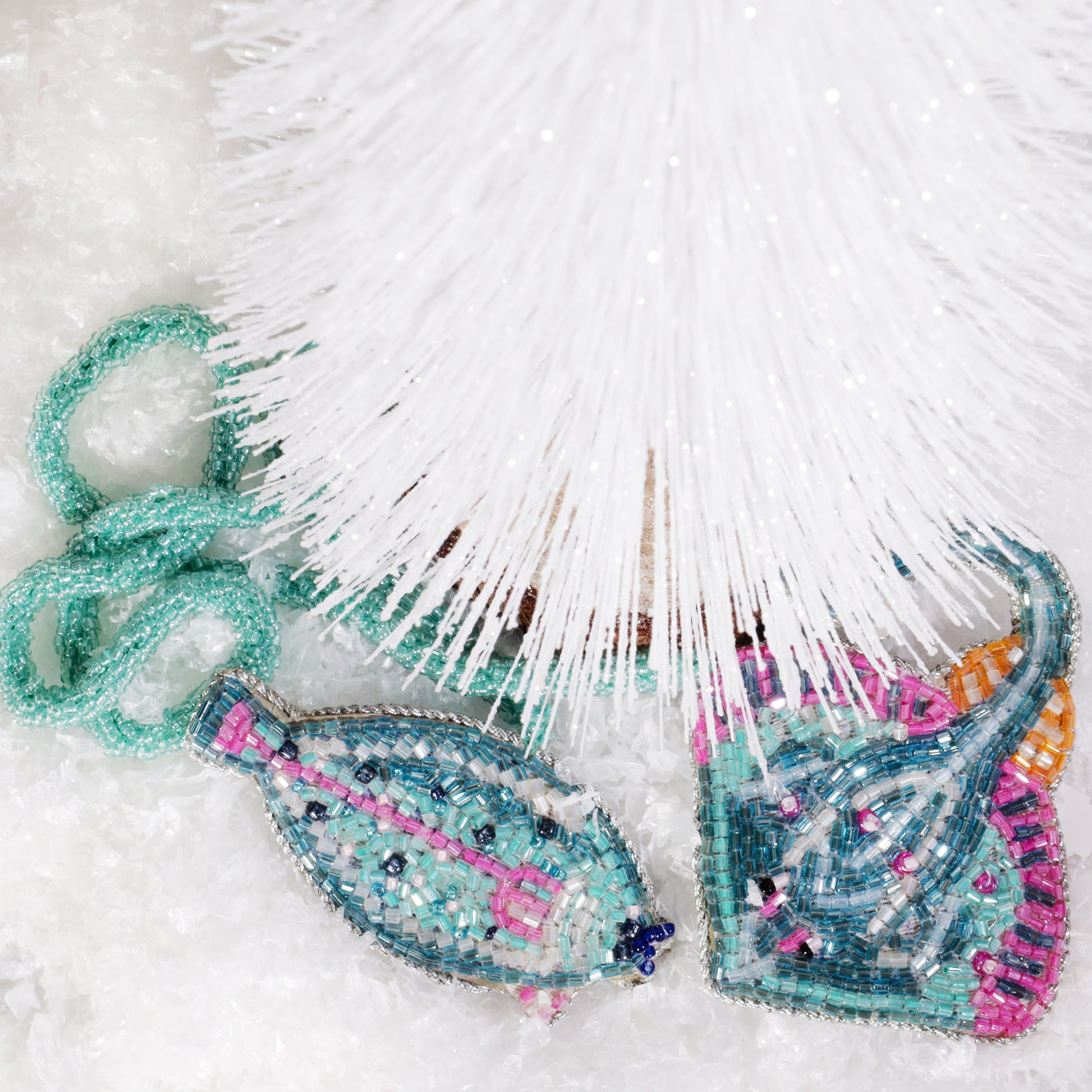 Two beaded ocean-themed bag charms, one shaped like a turquoise fish and the other like a stingray with pink and orange accents, displayed beneath a white Christmas tree on a bed of faux snow.