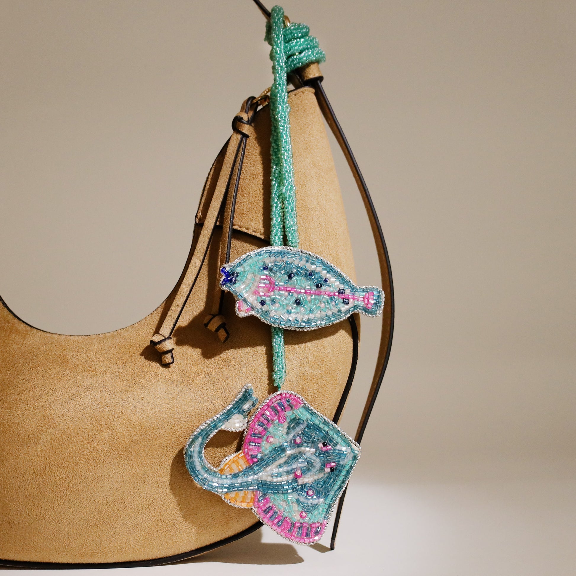 Tan suede handbag decorated with two beaded ocean-themed charms, one shaped like a turquoise fish and the other like a stingray, both featuring pink and blue detailing.