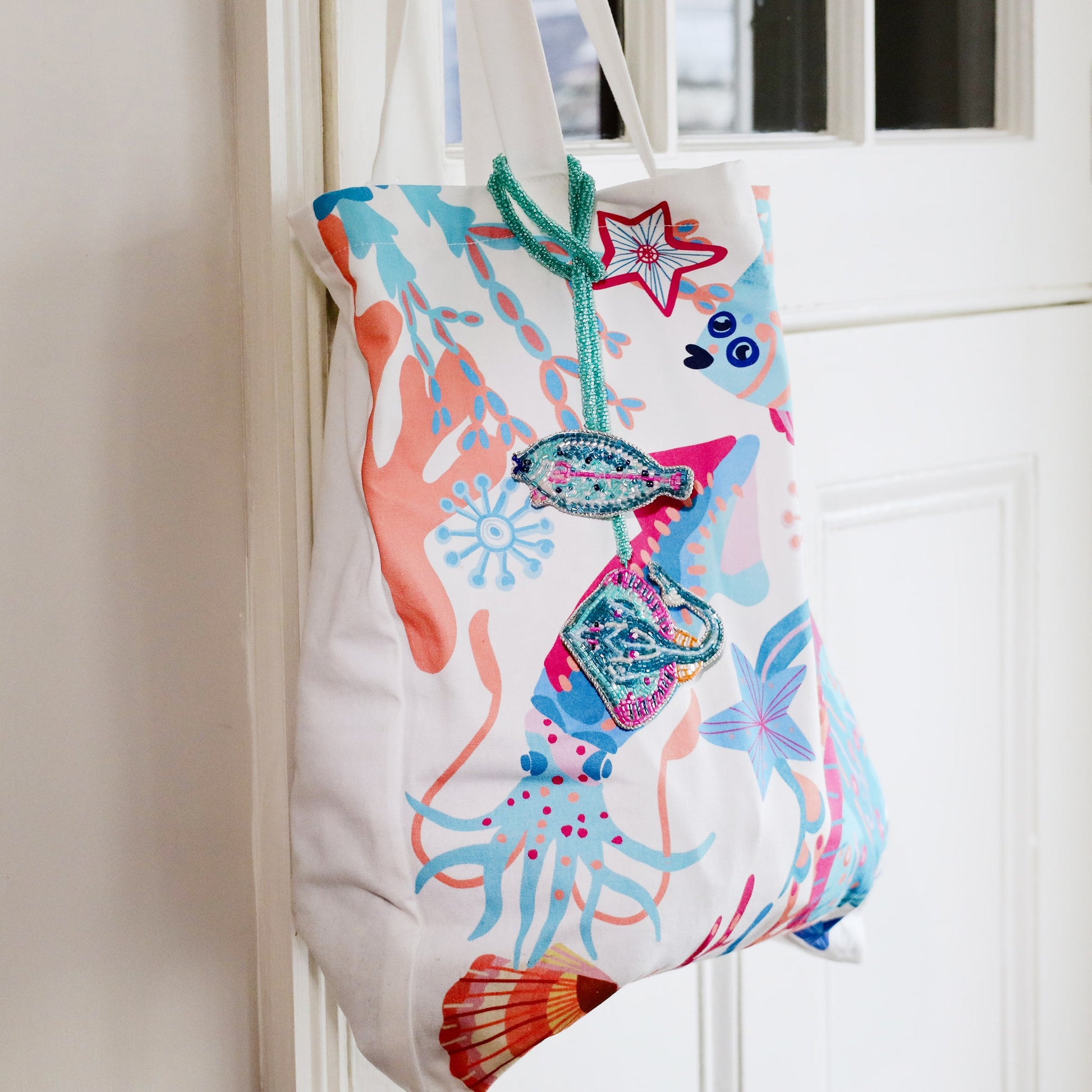 Colourful ocean-themed tote bag with coral and sea creature illustrations, decorated with two beaded bag charms shaped like a turquoise fish and a stingray with pink and blue detailing.