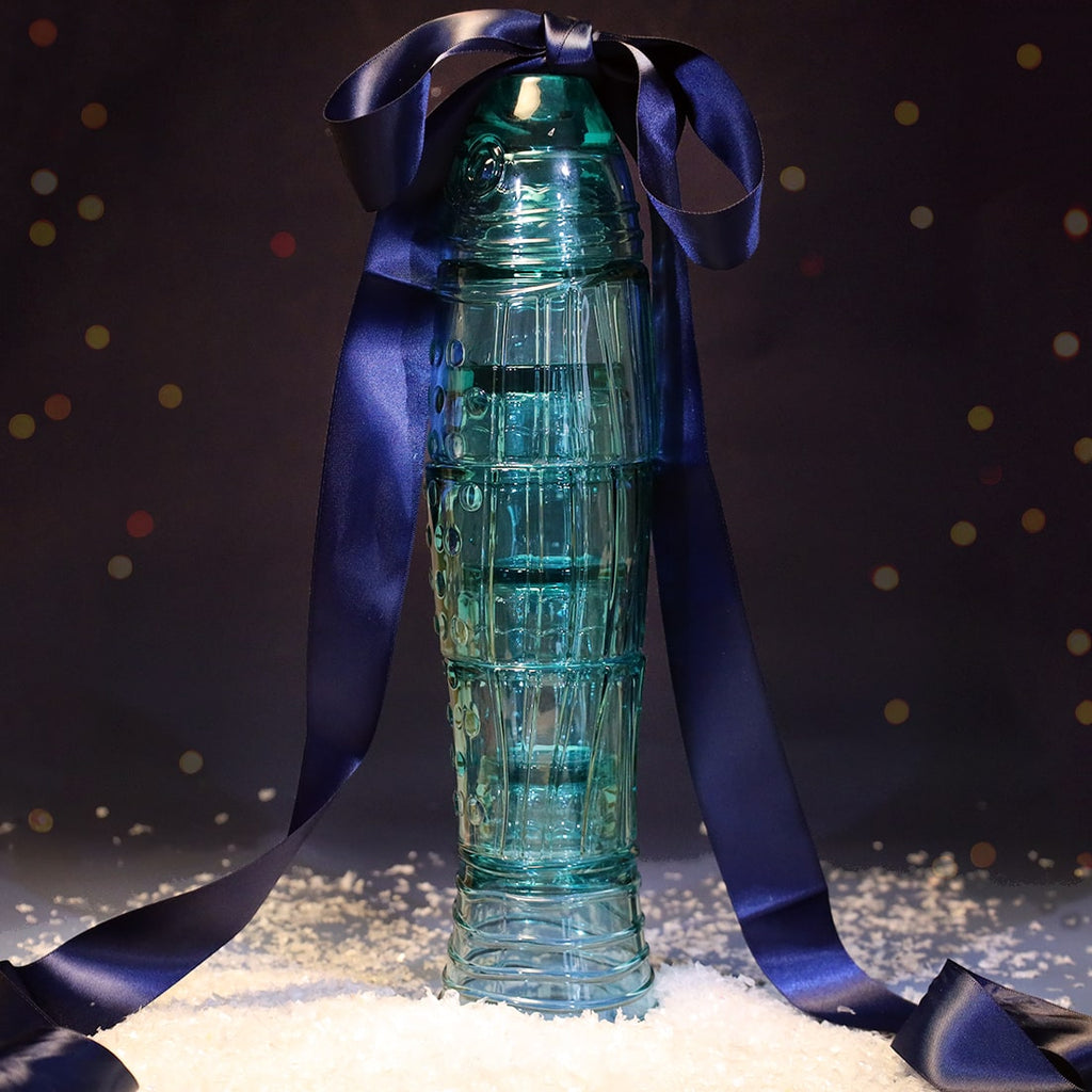Set of turquoise glass tumblers stacked in the shape of a fish, tied with a large navy satin ribbon and surrounded by faux snow against a softly lit festive background.