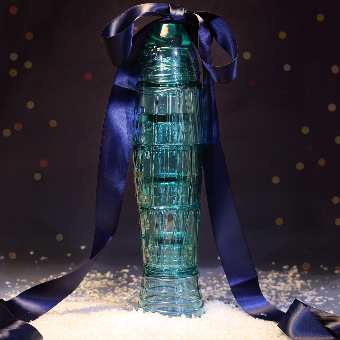 Set of turquoise glass tumblers stacked in the shape of a fish, tied with a large navy satin ribbon and surrounded by faux snow against a softly lit festive background.