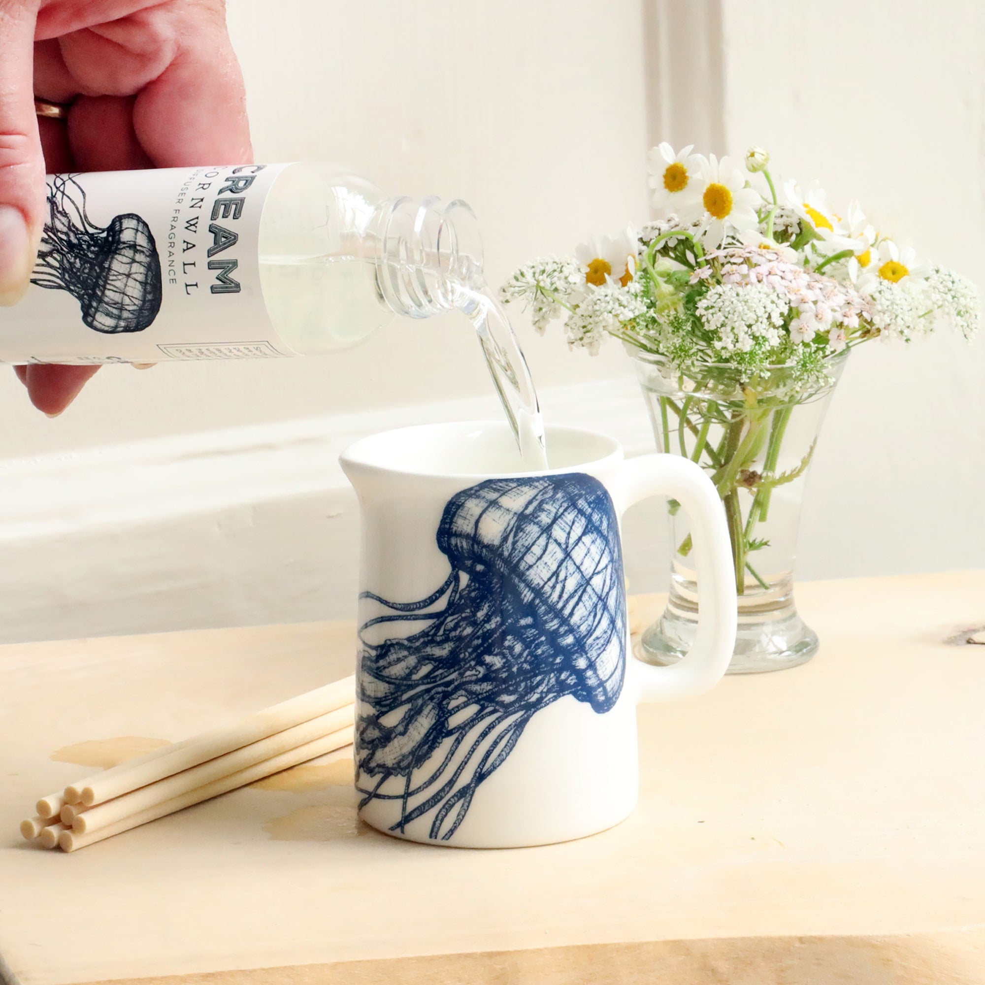 Mini Jug with jellyfish design on with reeds and small posy of flowers behind with a bottle of Fistral room diffuser oil being poured into the jug.