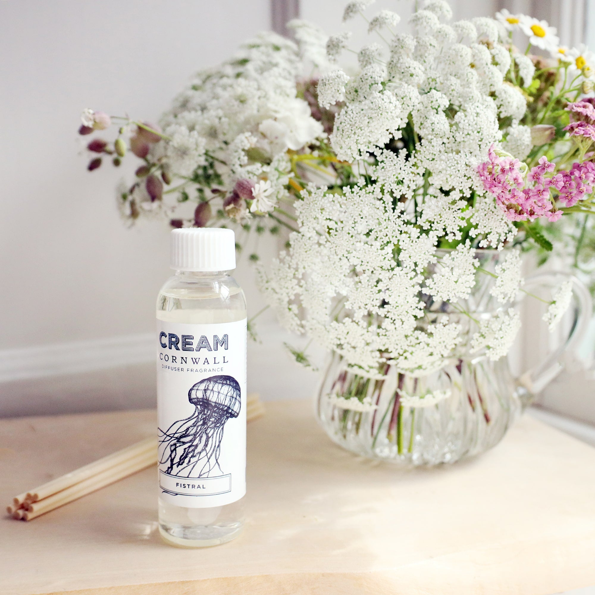 Bottle of Fistral room diffuser oil with octopus design on the label, sitting on a wooden block with the diffuser reeds laying jut to the side, and a vase of soft coloured flowers sitting behind.