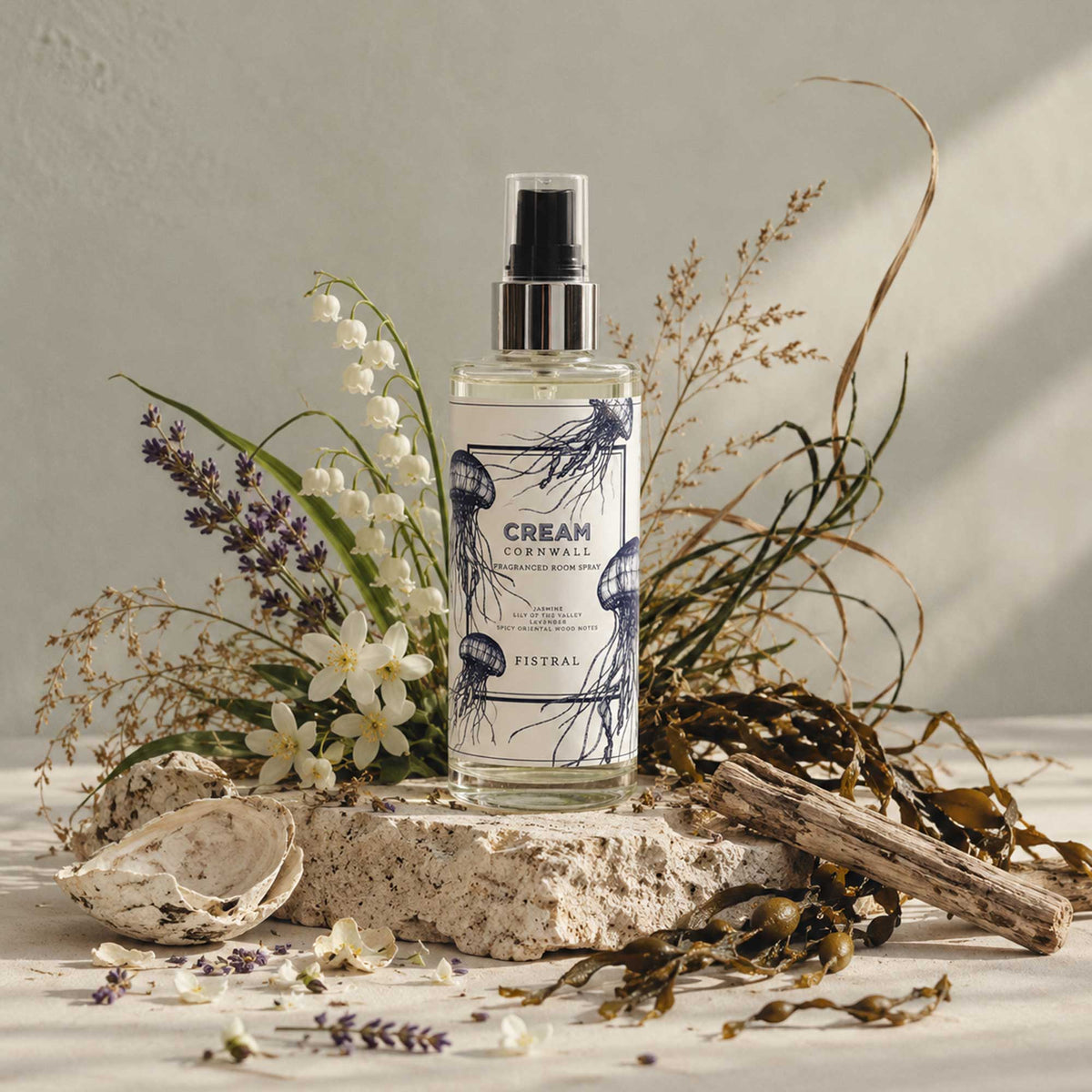 A bottle of Fistral Cream Cowwall Spray stands on a stone slab surrounded by dried flowers, seaweed, driftwood, and a shell in a styled studio setting.