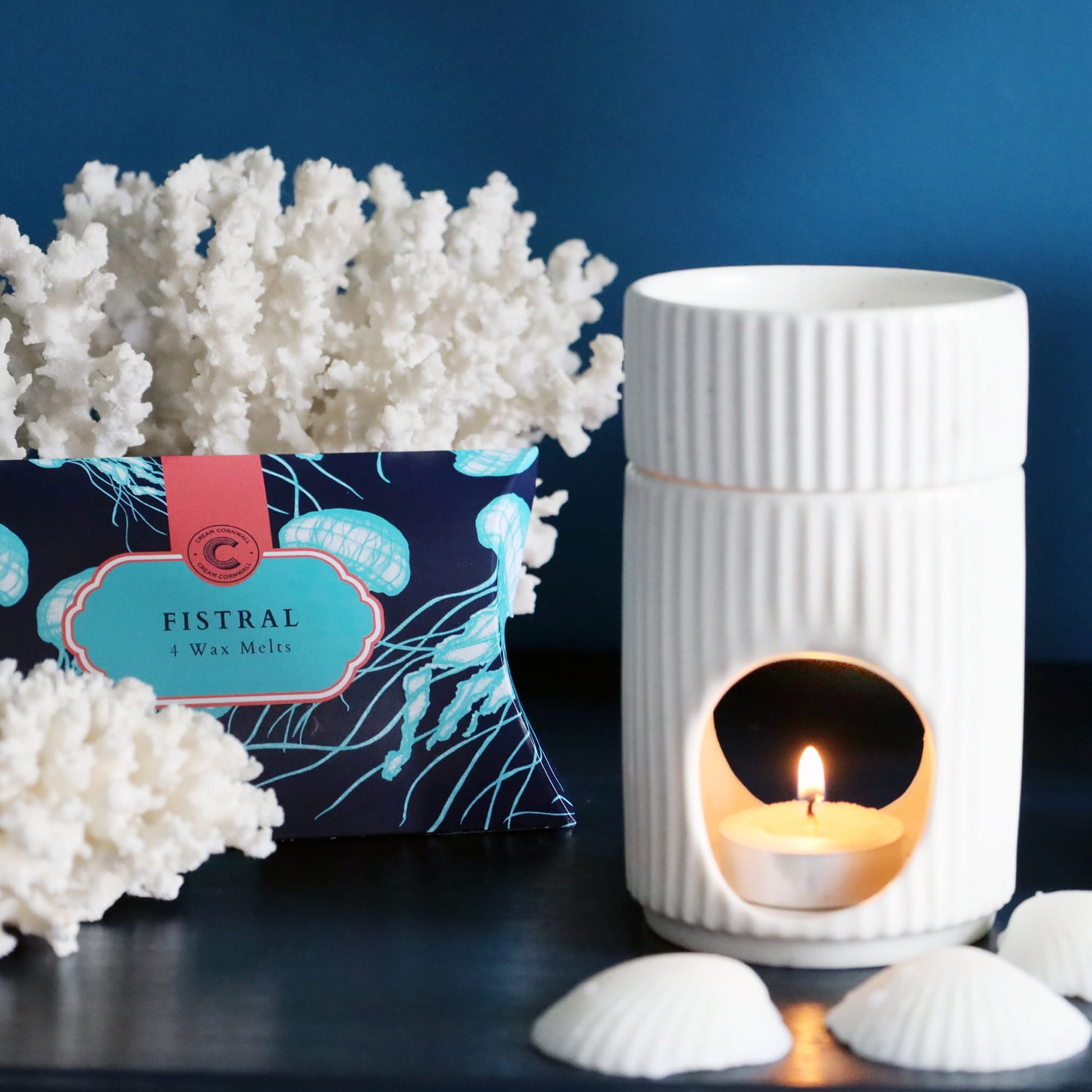 White textured wax melter with a lit candle, next to a box of FISTRAL wax melts on a dark surface with white coral and shells.