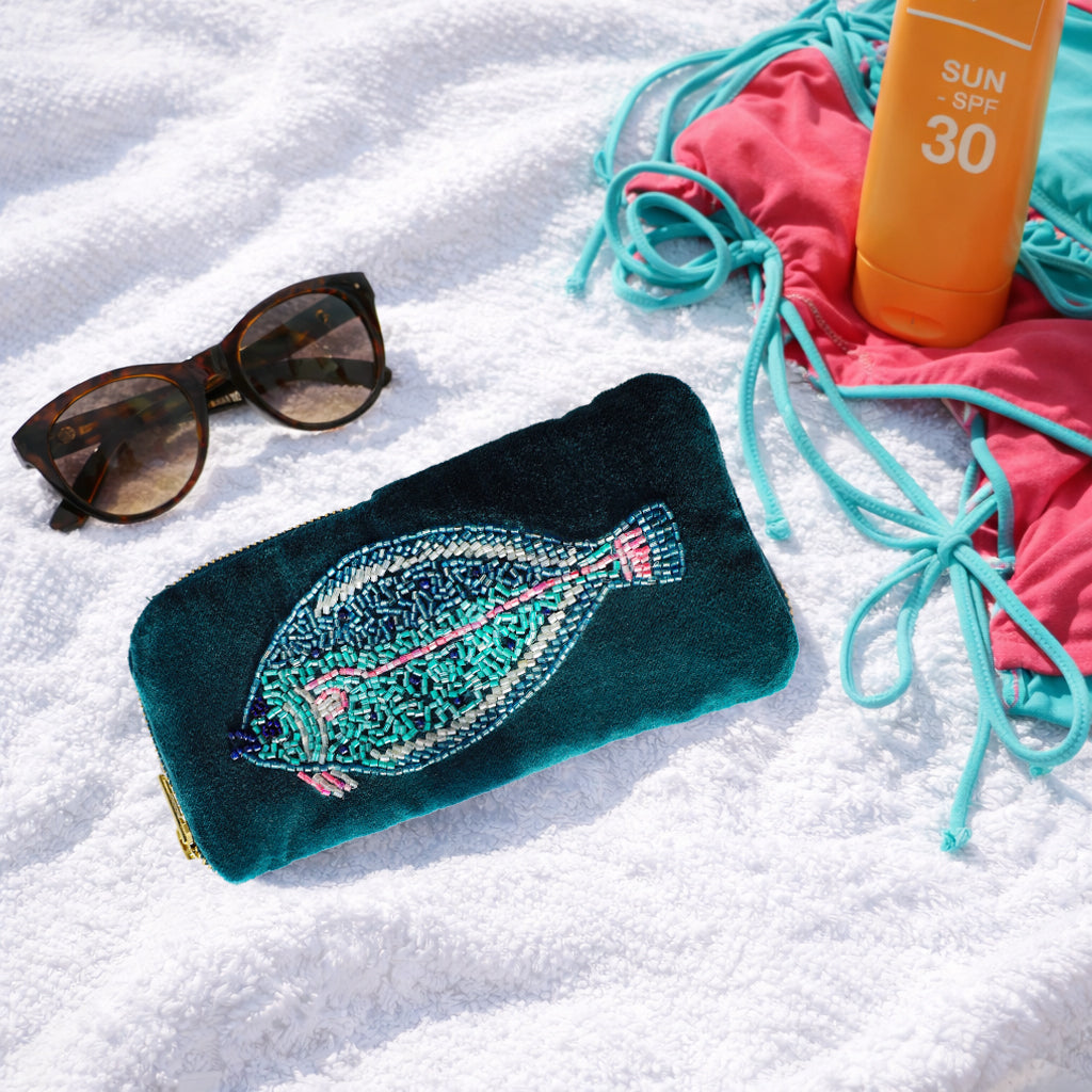 Teal pouch with fish design, sunglasses, and sunscreen on a sandy surface