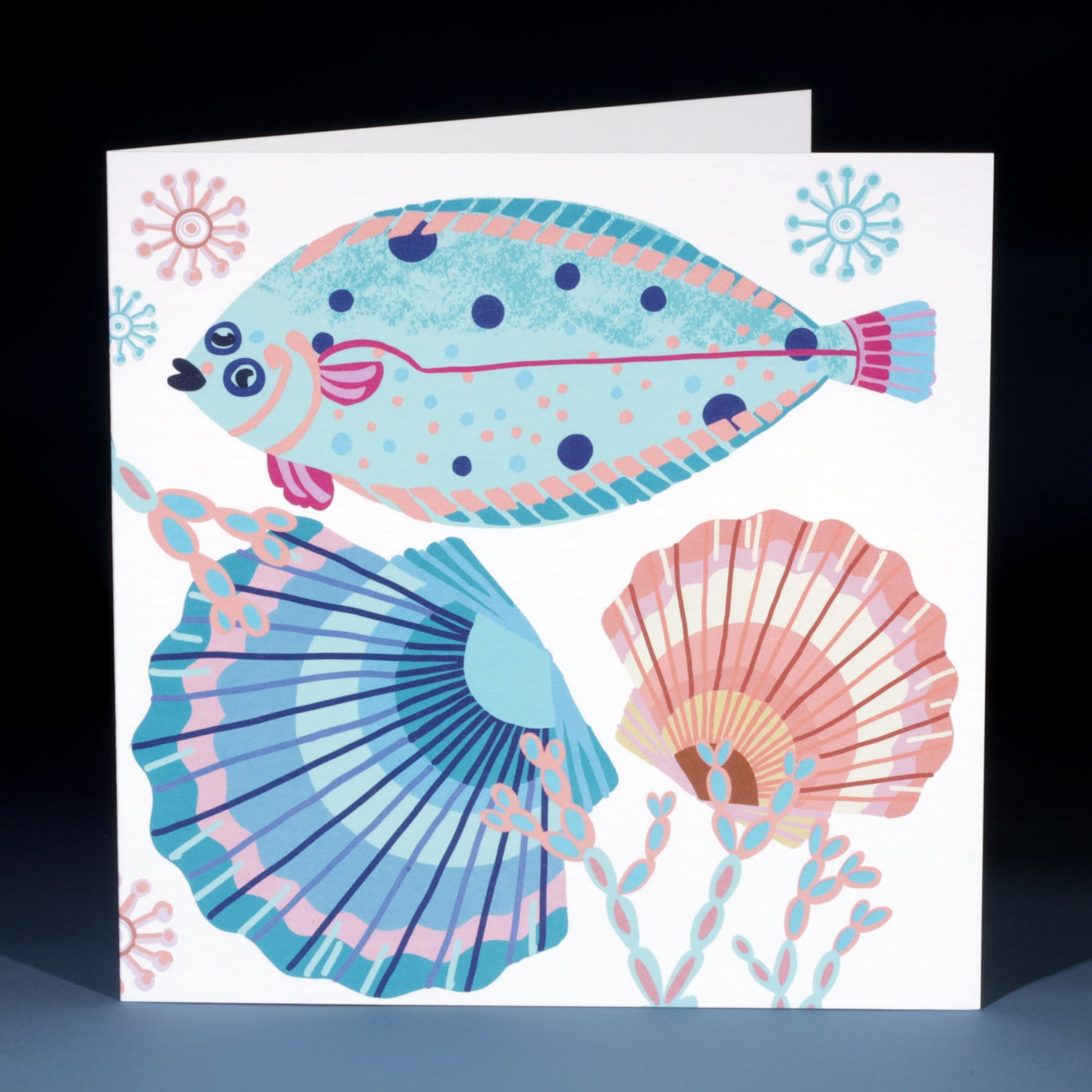 Greeting card featuring a colourful illustration of a flat fish with vibrant blue, pink, and purple patterns, surrounded by two decorative scallop shells and abstract sea plants on a white background.