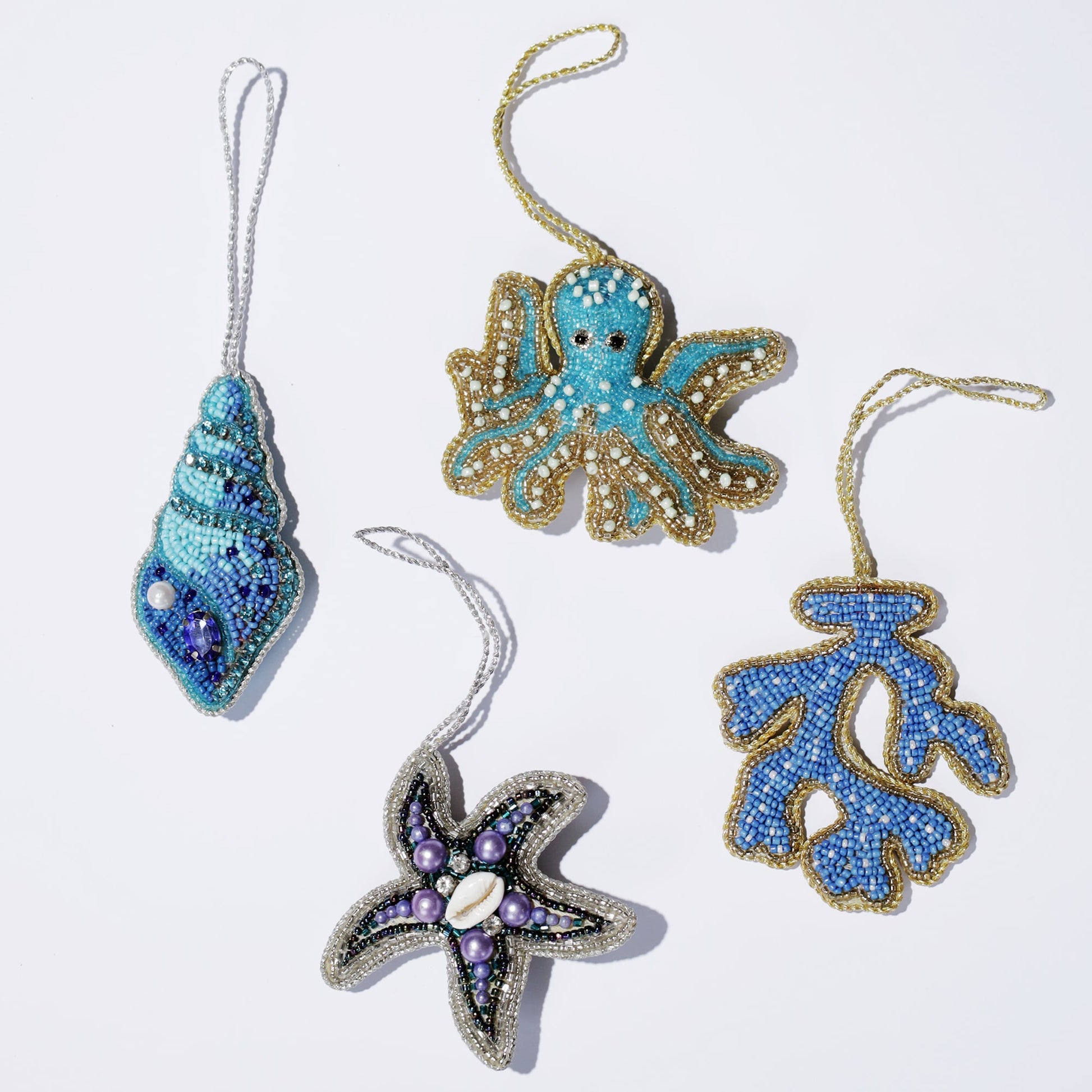 Flat lay of four ocean-inspired beaded ornaments on a white background, including a blue seashell with pearl details, a turquoise octopus outlined in gold, a blue coral branch with gold trim, and a starfish adorned with purple beads and a central white shell. The intricate beadwork and vibrant colors evoke a coastal theme.