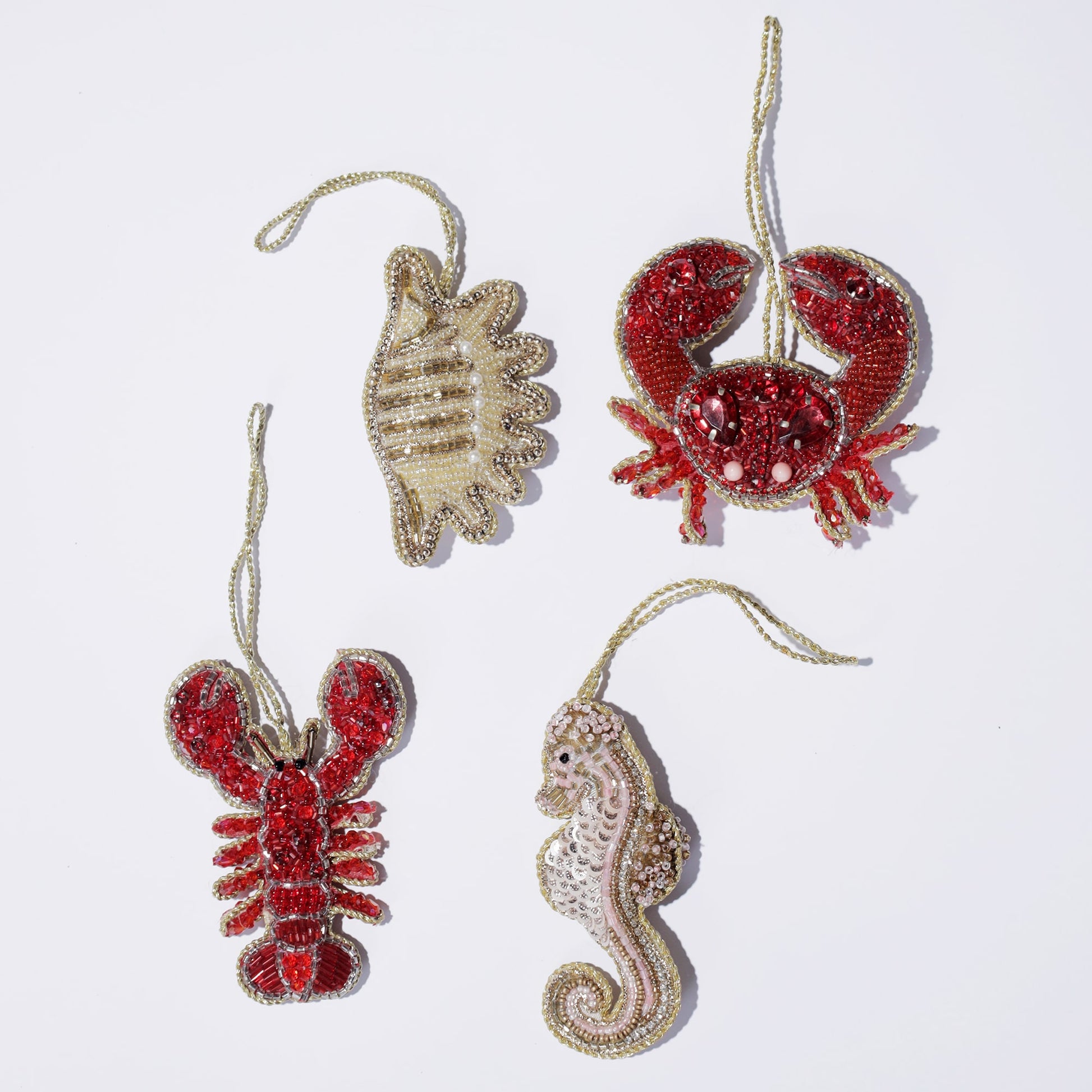 Flat lay of four ocean-themed beaded ornaments on a white background, featuring a shimmering gold seashell, a red crab with jeweled eyes, a vibrant red lobster, and a pink seahorse adorned with gold accents. Each ornament is intricately designed, evoking coastal charm and marine life.