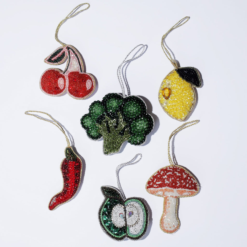 Flat lay of six beaded ornaments on a white background, each depicting a different fruit or vegetable: bright red cherries, a yellow lemon with a green leaf, a red chili pepper, a green broccoli floret, a halved green apple, and a red and white mushroom. Each piece is intricately crafted with colorful beads, adding a playful and vibrant touch to the collection.
