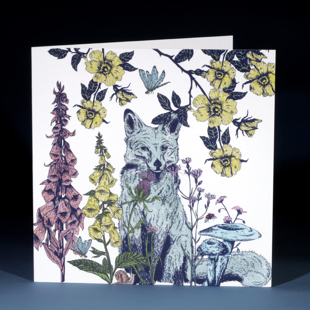 Front of a greeting card featuring an intricately illustrated fox surrounded by wildflowers, mushrooms, and plants in soft tones of yellow, pink, blue, and green on a white background. A butterfly and ladybird add whimsical details to the woodland scene.