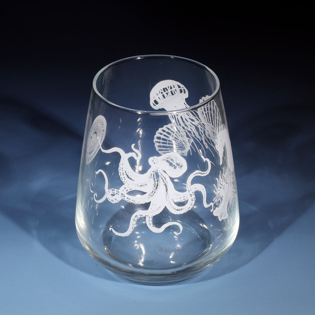 The image shows a clear glass with intricate white prints of underwater sea creatures, including an octopus, jellyfish, seahorse and other marine life. The artwork is detailed and wraps around the glass, creating an aquatic-themed design. The glass is placed on a smooth blue surface, with soft lighting casting shadows and highlighting the delicate etchings.