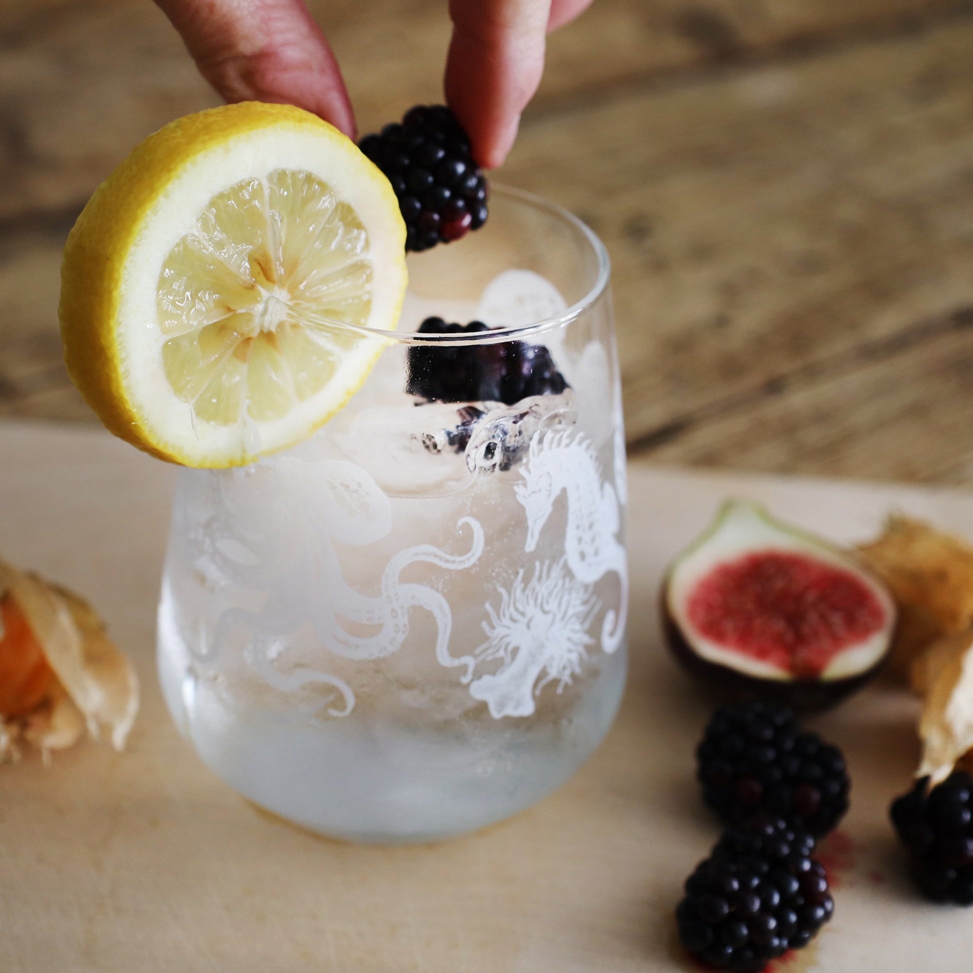 
A close-up of a glass with an intricate marine design featuring an octopus, seahorse, and seaweed, filled with a clear, icy beverage. A lemon slice is perched on the rim, and a hand is gently placing a blackberry into the glass, which already contains several other blackberries. In the background, there are additional blackberries, a half fig, and a cape gooseberry, all resting on a wooden surface, adding a rustic and fresh feel to the image.