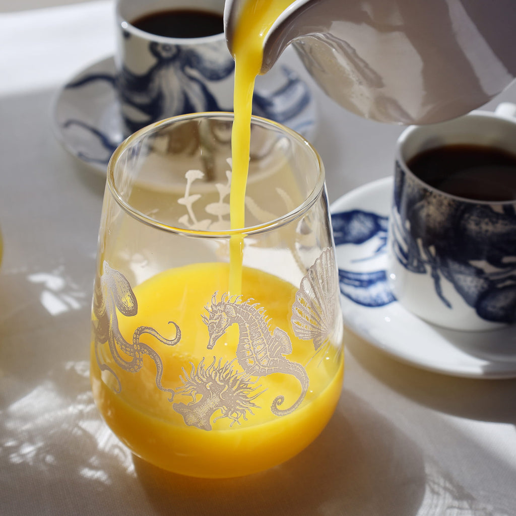 Orange juice being poured into a glass with sea creature designs, beside blue-and-white coffee cups on a sunlit table.