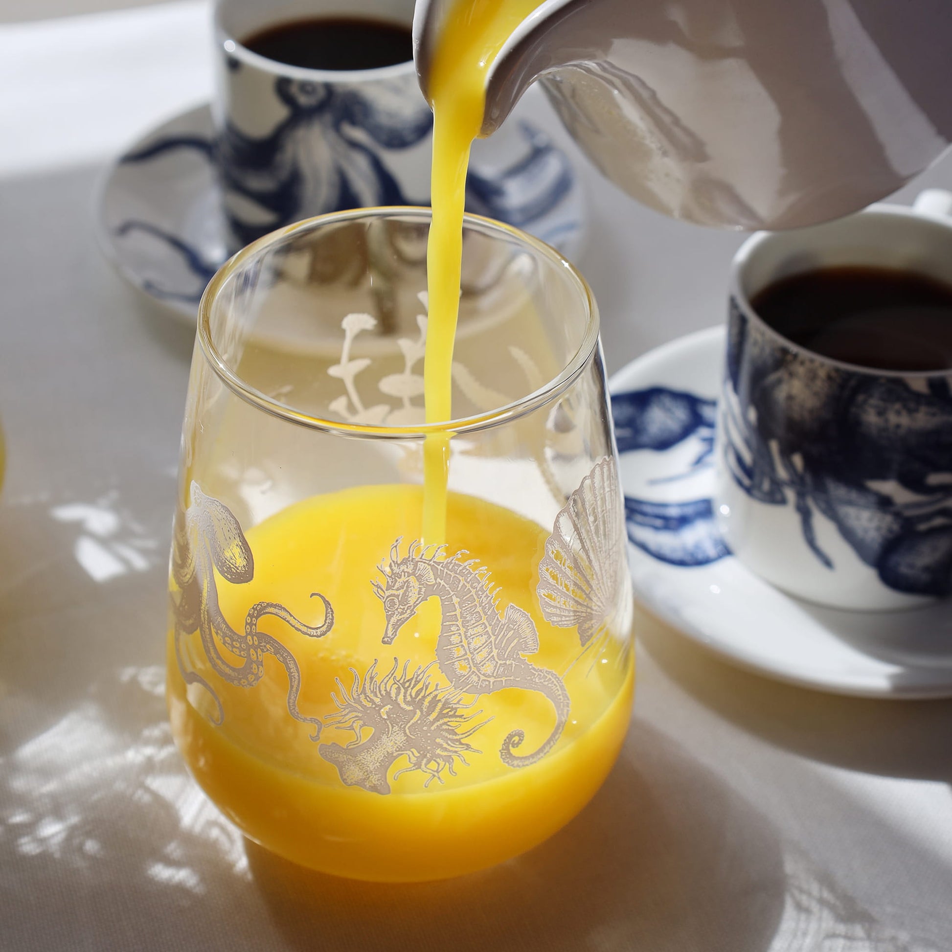 Orange juice being poured into a glass with sea creature designs, beside blue-and-white coffee cups on a sunlit table.