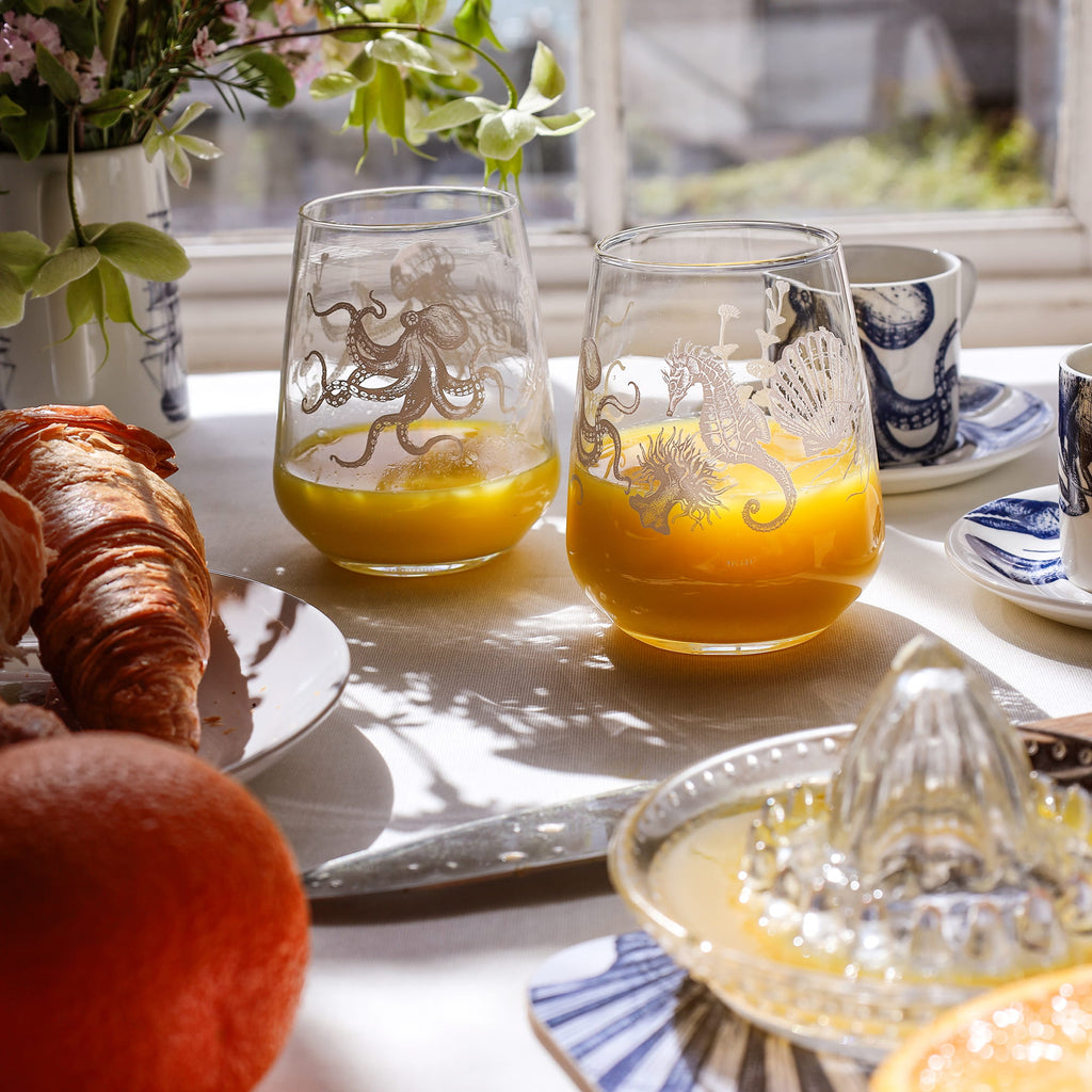 Sunlit breakfast table with two glasses of orange juice featuring sea creature designs, croissants, and coffee by a window.