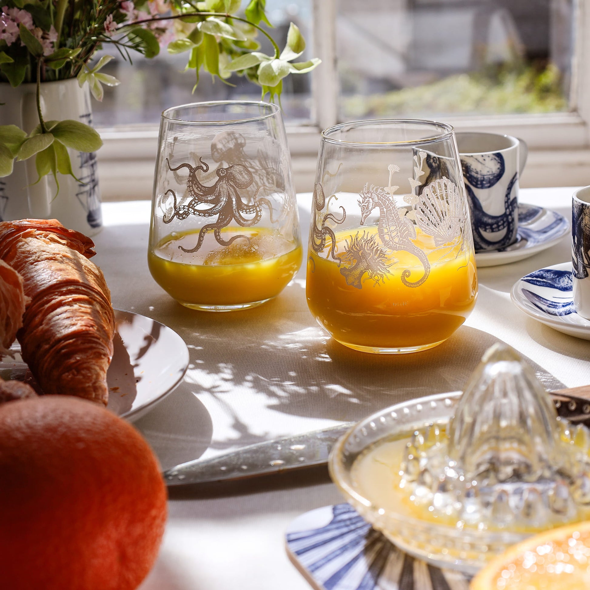 Sunlit breakfast table with two glasses of orange juice featuring sea creature designs, croissants, and coffee by a window.