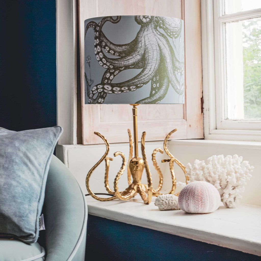 A table lamp with a gray octopus-patterned drum shade and a gold tentacle-shaped base, styled on a windowsill with coral and seashells.