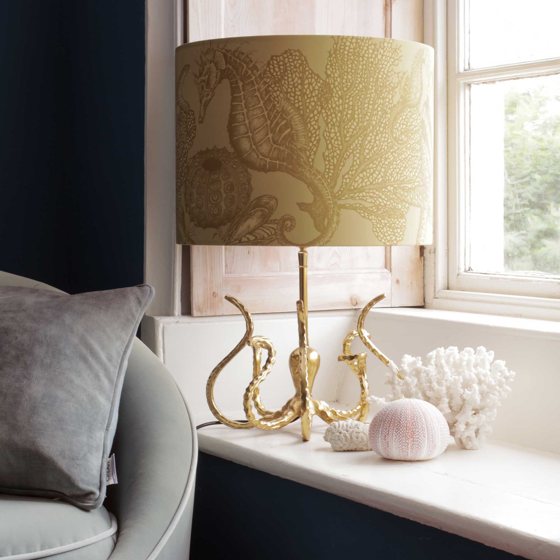 A table lamp with a beige and gold seahorse and coral patterned drum shade and a sculptural gold octopus base, styled on a white windowsill next to a gray armchair.