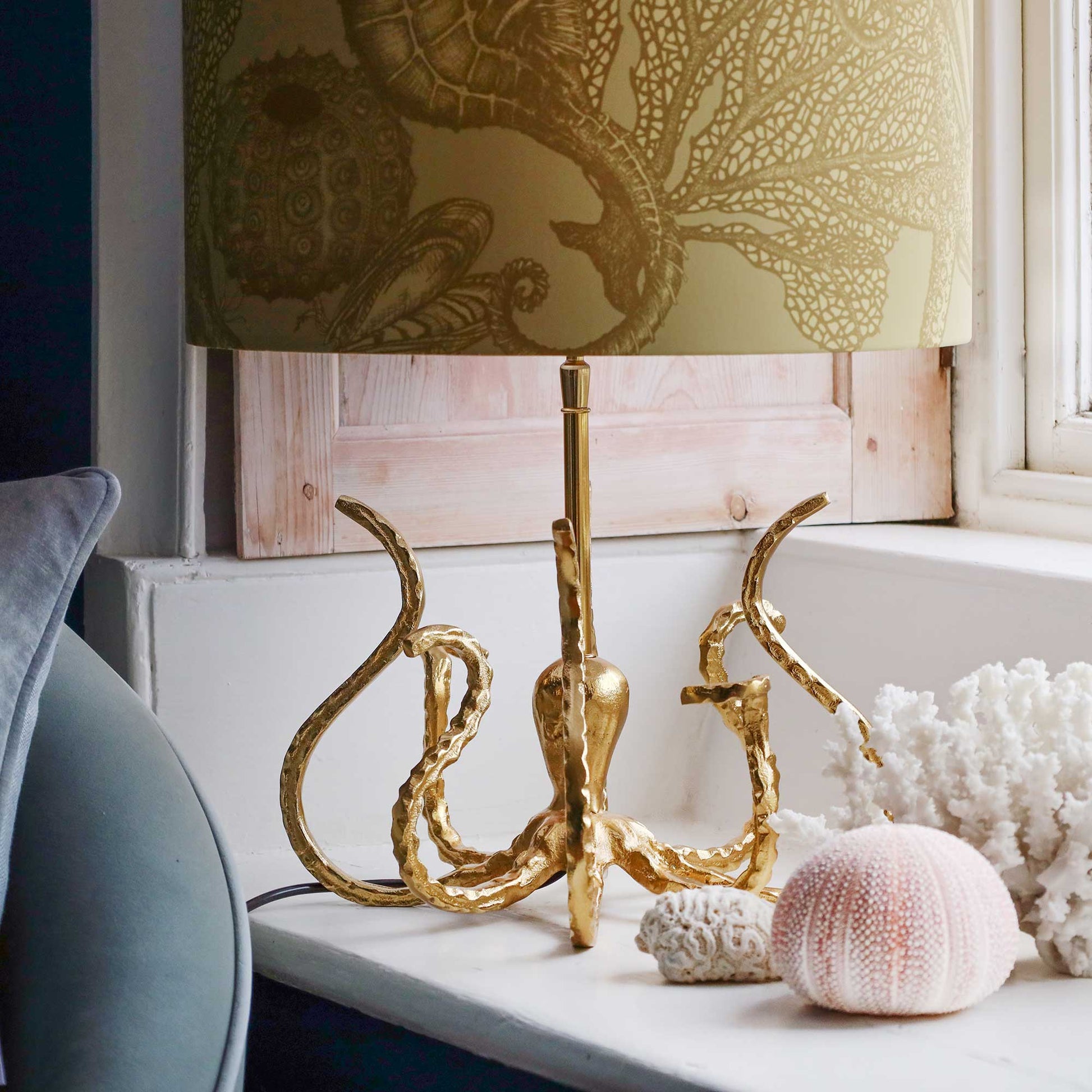 A table lamp with a gold octopus-shaped base and a beige patterned shade, styled on a windowsill with seashells and coral.
