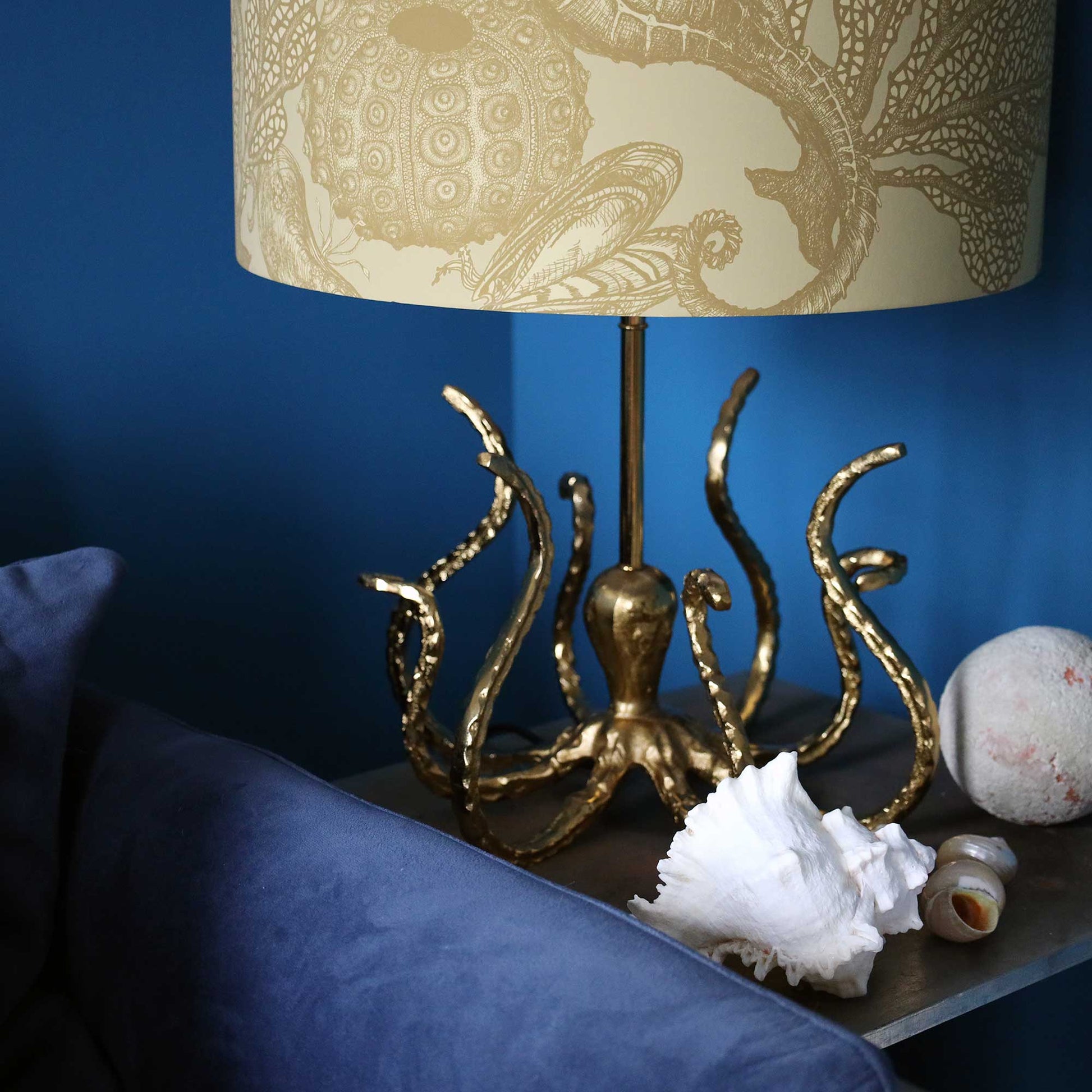 A table lamp with a gold octopus-shaped base and a beige patterned drum shade, styled on a side table with seashells against a deep blue wall.