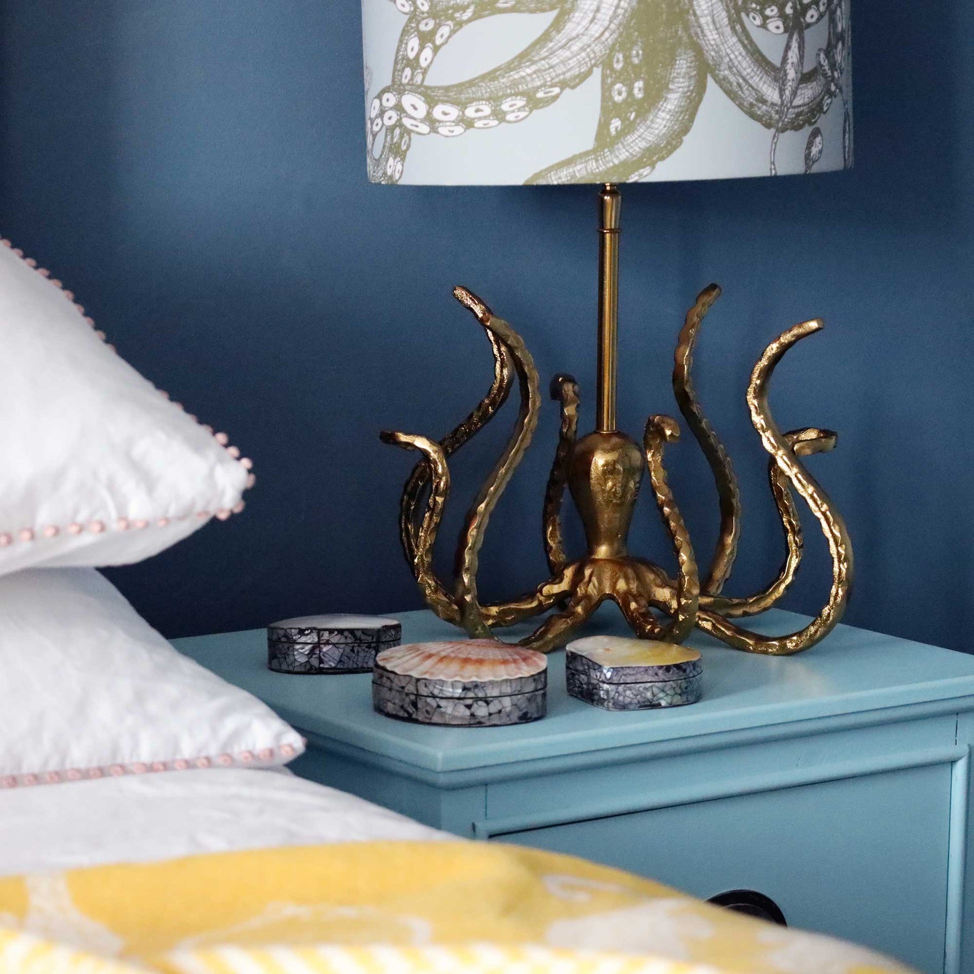 A brass octopus-shaped table lamp with a white and olive-green octopus patterned shade, placed on a light blue nightstand next to a bed with white pillows and a yellow blanket.