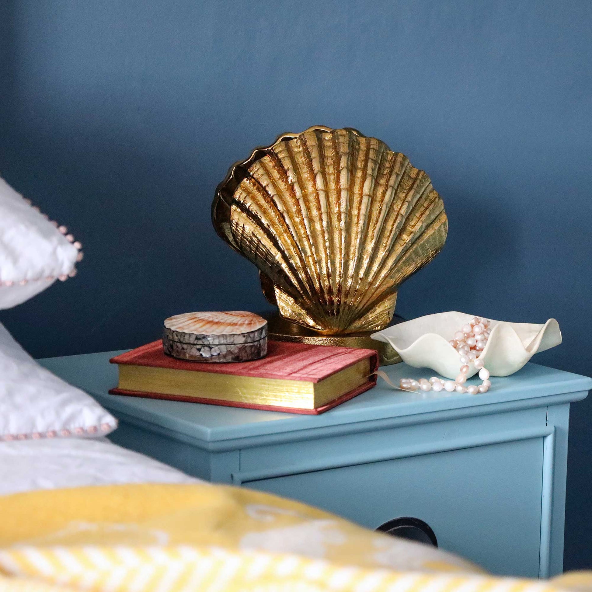 A decorative gold-toned scallop shell lamp base displayed on a light blue bedside table, styled with a pink book, a small round box, a white shell dish with pearls, and part of a bed in a bedroom setting.