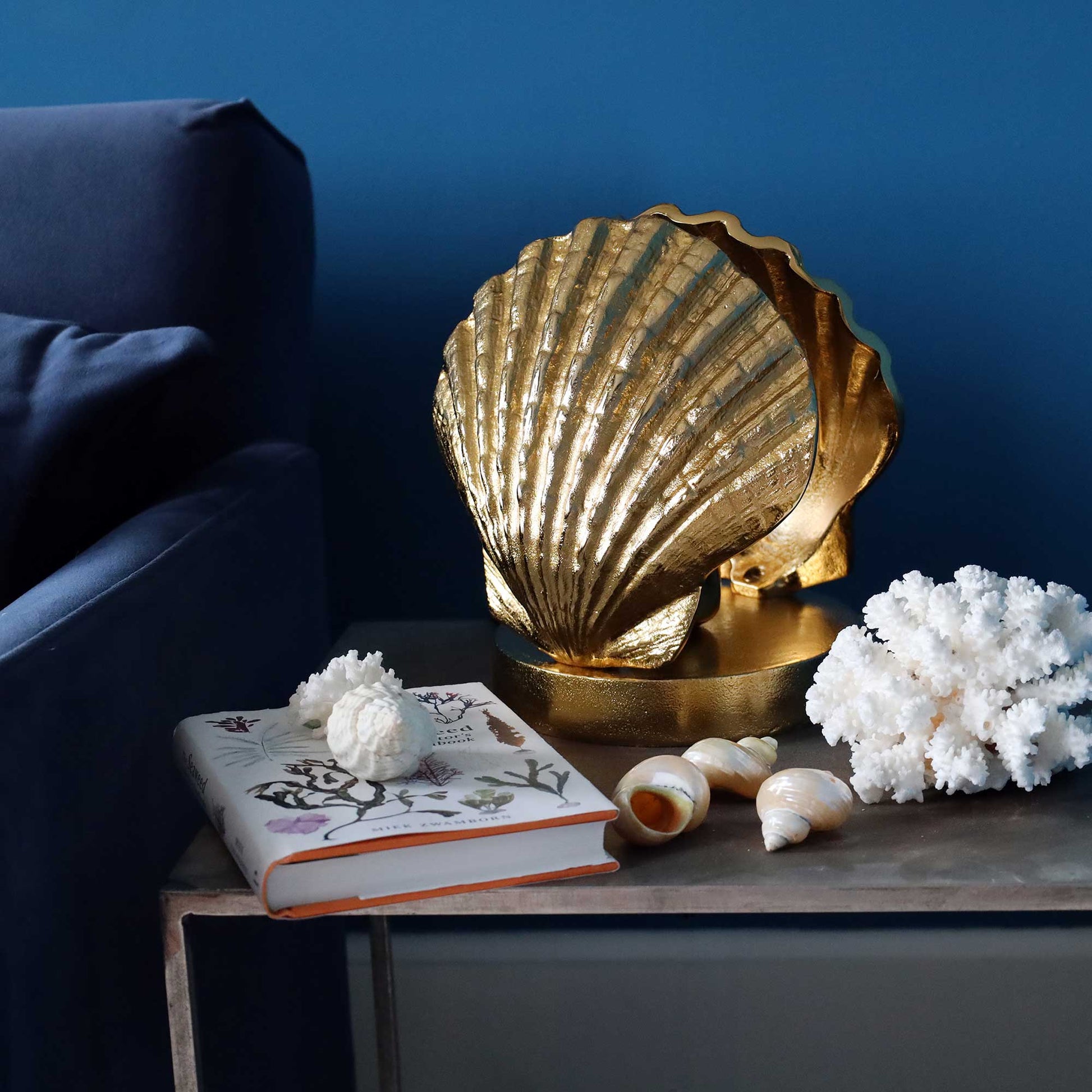 A decorative gold shell lamp shaped like a scallop shell sits on a dark table next to a navy blue armchair, with white coral and seashells around it.