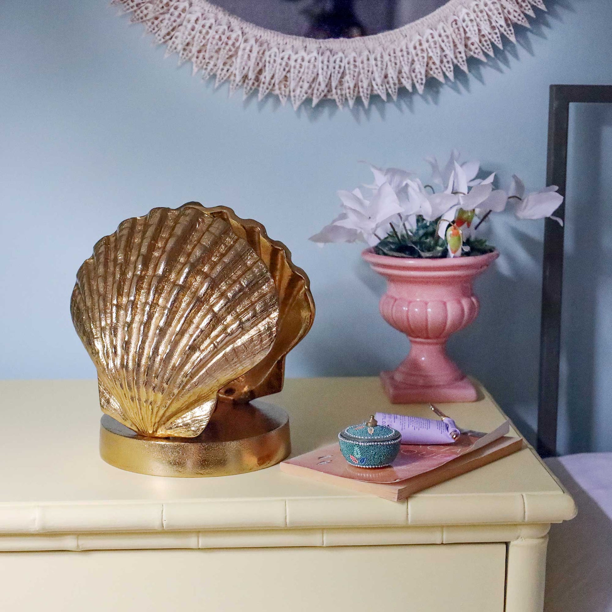 A large metallic gold scallop shell lamp on a round base, displayed on a yellow-coloured nightstand next to a pink flower pot and books against a light blue wall.