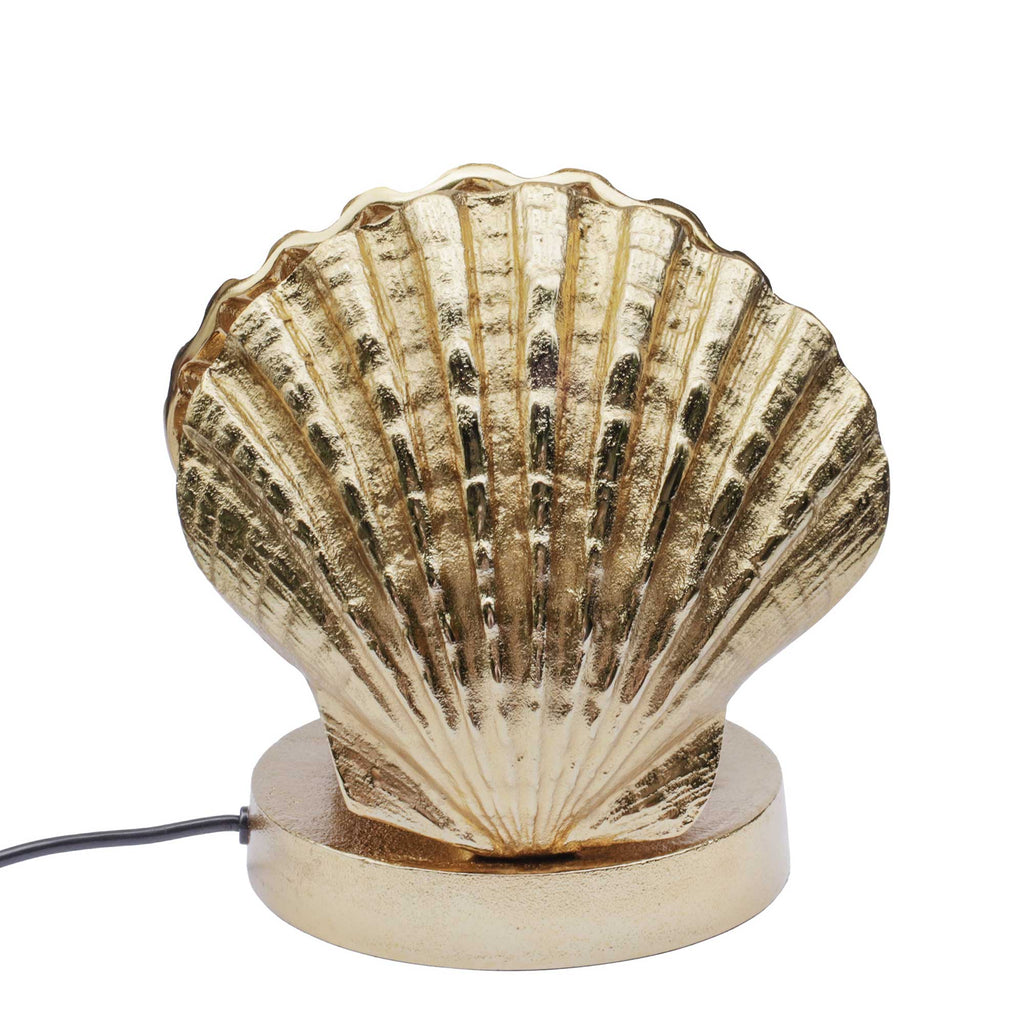 A decorative table lamp shaped like a metallic gold scallop shell on a round base, with a black power cord visible on the left, set against a plain white background.