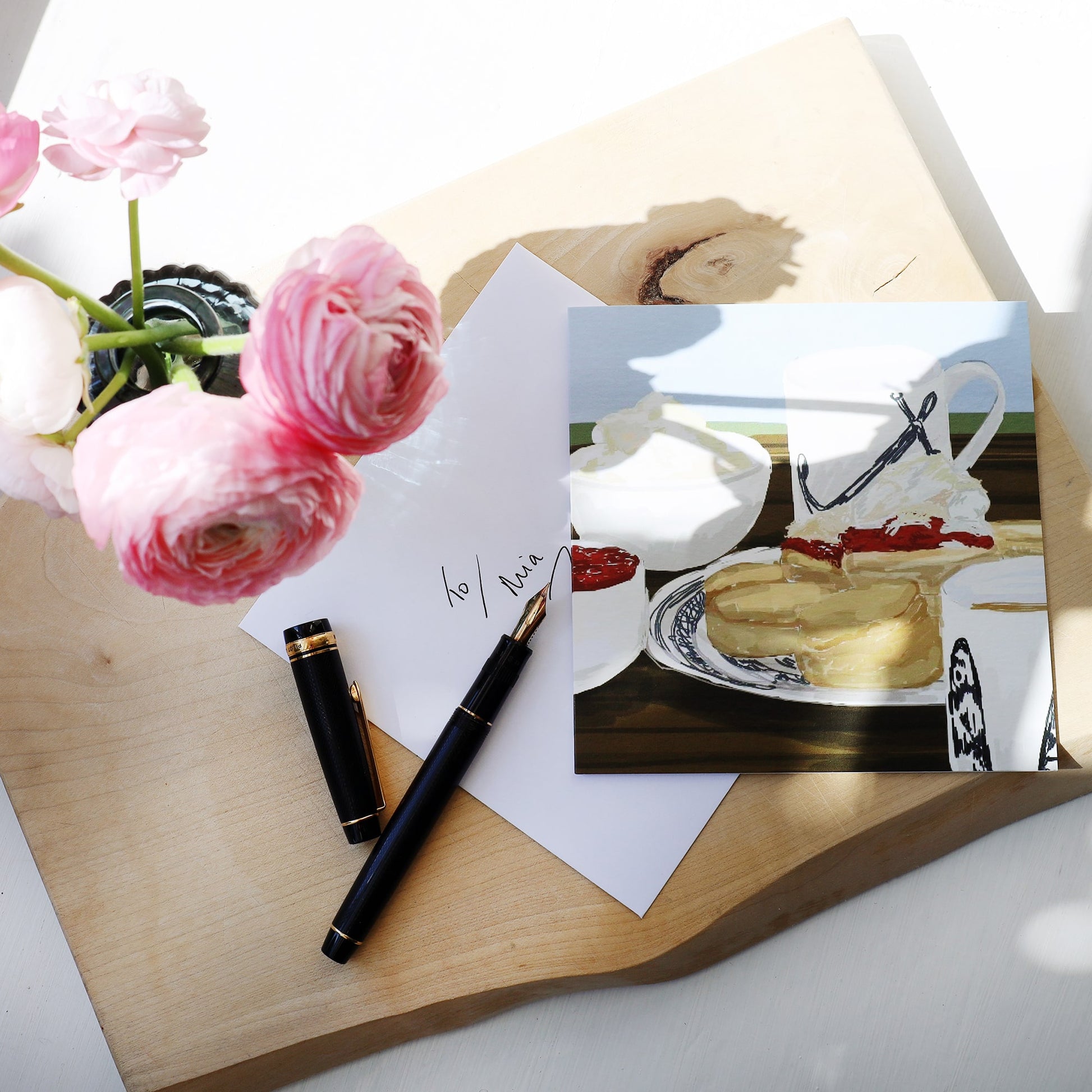 Greeting card featuring a cream tea illustration with scones, jam, clotted cream, and a mug, placed next to an envelope handwritten “To Mia,” a black fountain pen, and a vase of pink ranunculus flowers on a light wooden board.