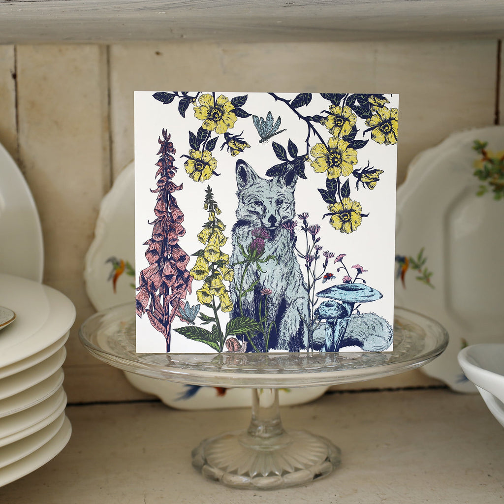 Greeting card featuring an illustrated fox surrounded by detailed botanical elements including foxgloves, yellow wildflowers, mushrooms, and a butterfly. The card is displayed standing on a glass cake stand in a vintage-style cupboard with stacked white plates and floral china.