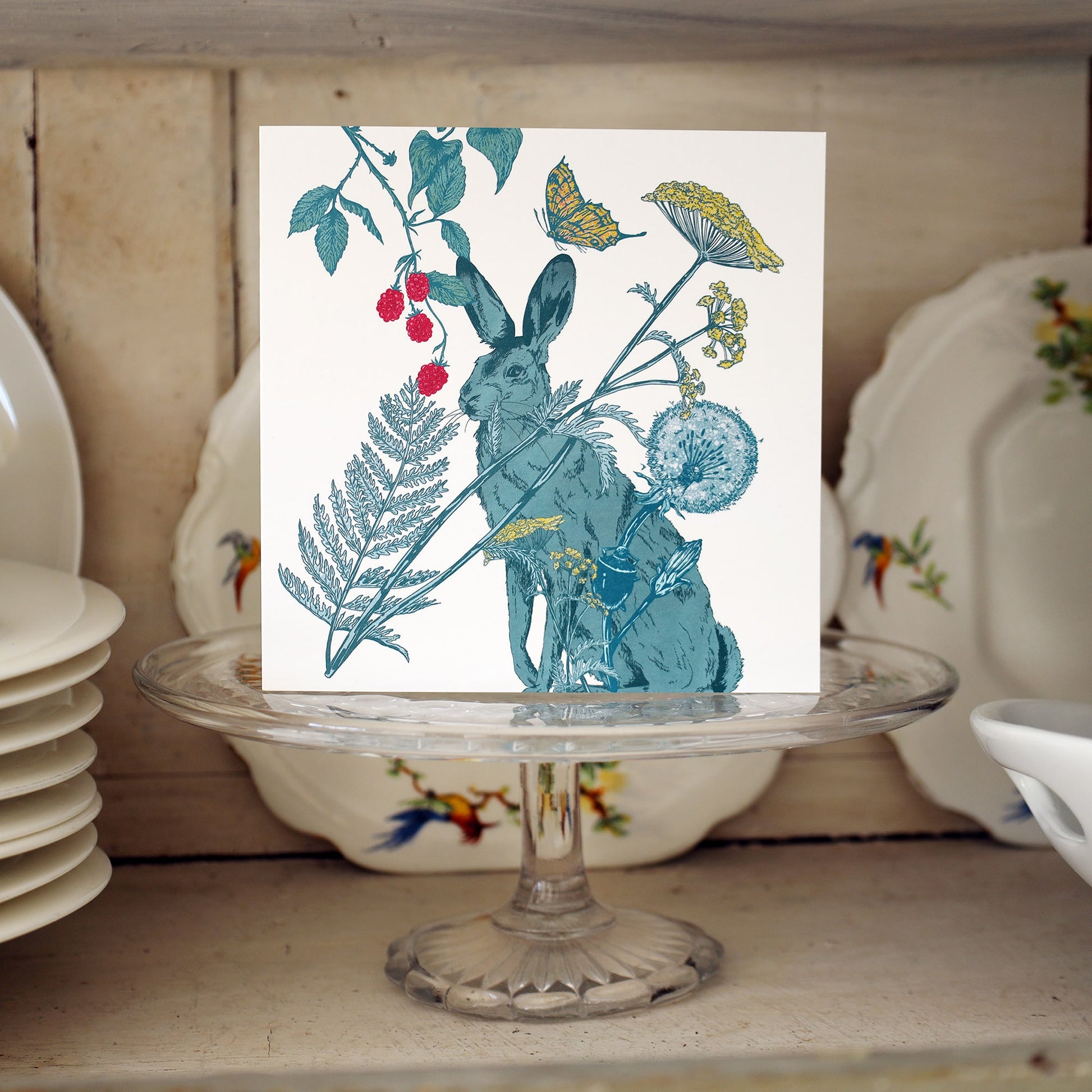 Greeting card featuring a detailed illustration of a hare surrounded by botanical elements including berries, wildflowers, ferns, and a butterfly. The card is displayed upright on a glass cake stand in front of vintage floral plates on a wooden shelf.
