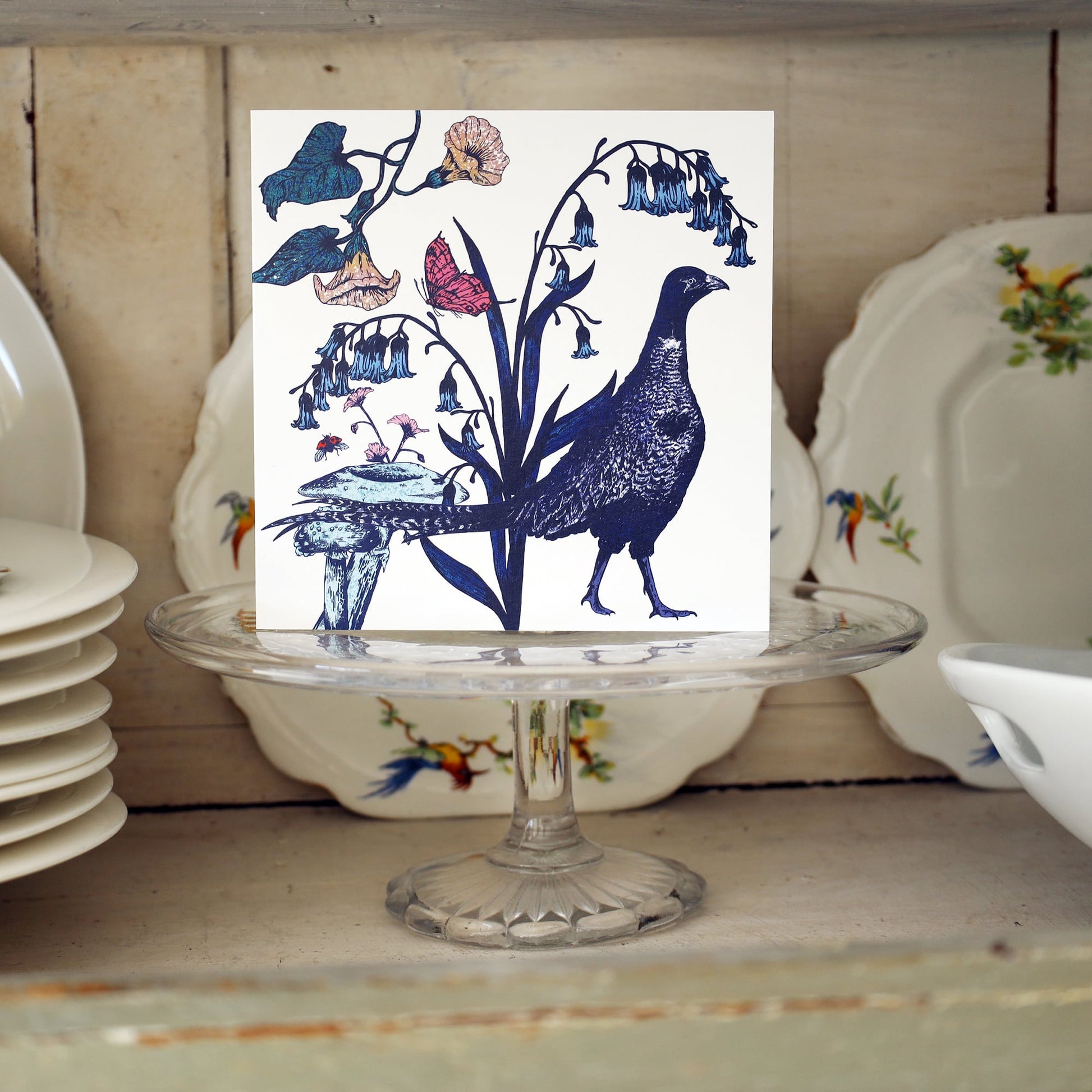 Greeting card featuring an illustrated pheasant surrounded by bluebells, mushrooms, butterflies, and wildflowers, displayed upright on a glass cake stand in front of vintage floral china plates on a wooden shelf.