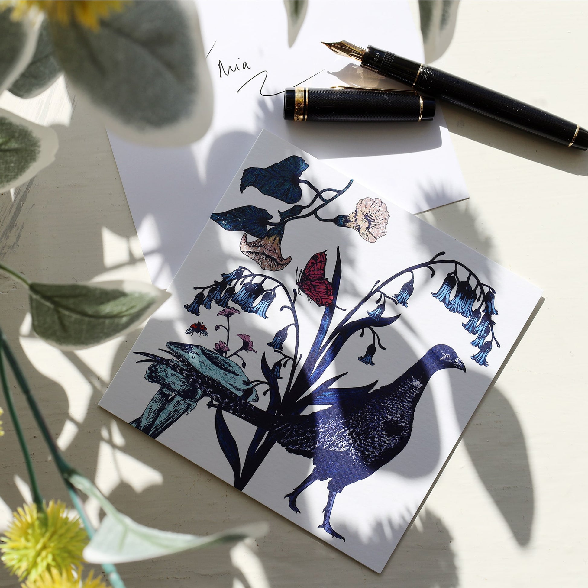 Illustrated pheasant greeting card lying on a white surface next to an envelope with "To Mia" handwritten on it and a black and gold fountain pen, with soft floral shadows cast across the scene.