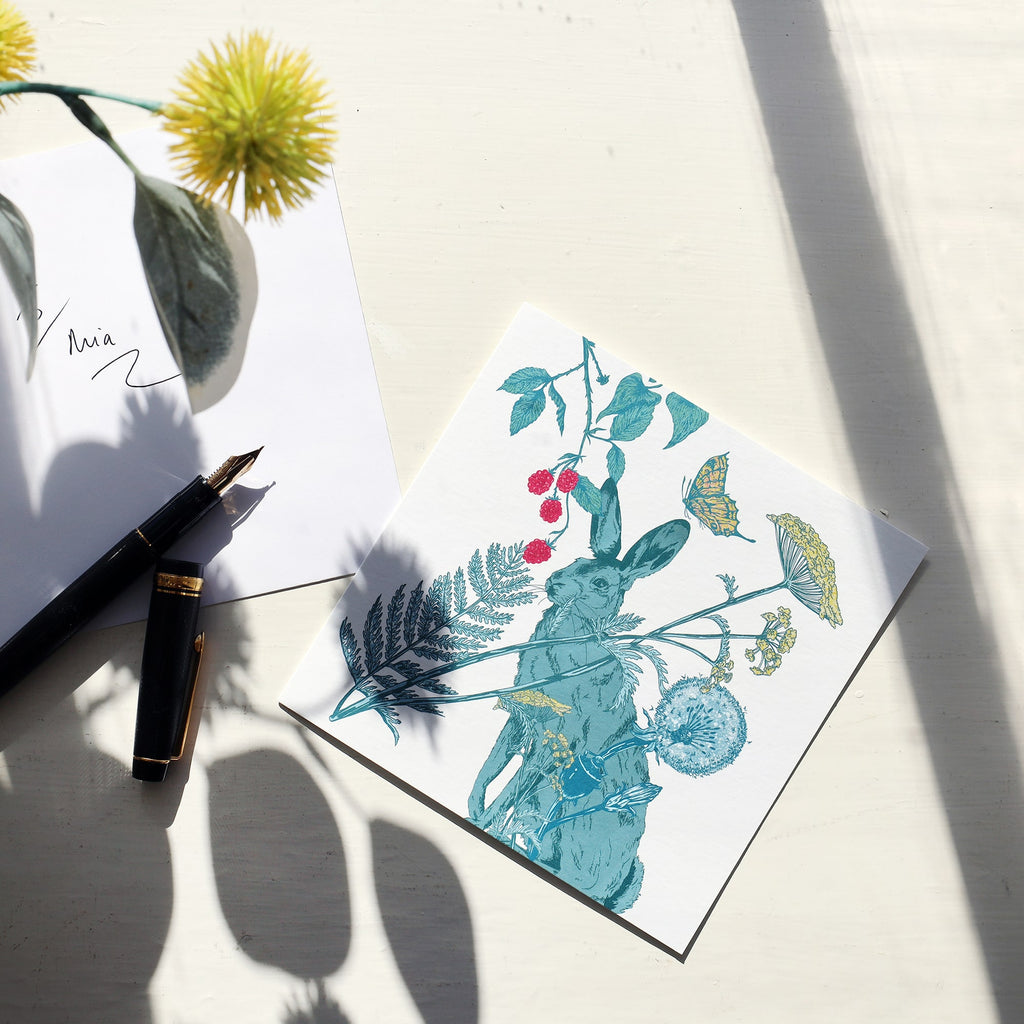Greeting card featuring an illustrated hare surrounded by foliage, berries, wildflowers, and a butterfly, resting on a sunlit white surface. Beside it is an envelope with handwritten text “To Mia,” a black fountain pen with gold details, and the shadow of a yellow flower cast over the scene.