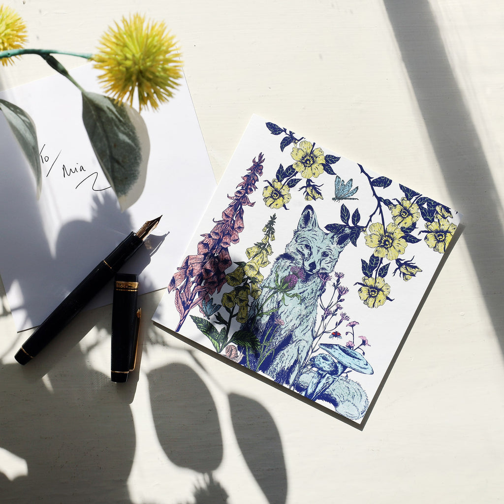 A greeting card with an illustrated fox surrounded by yellow flowers, mushrooms, and wild plants is placed on a white surface. Next to it is an envelope addressed "To Mia" and a black fountain pen. Soft sunlight and shadows from nearby flowers add a warm, natural ambiance to the scene.
