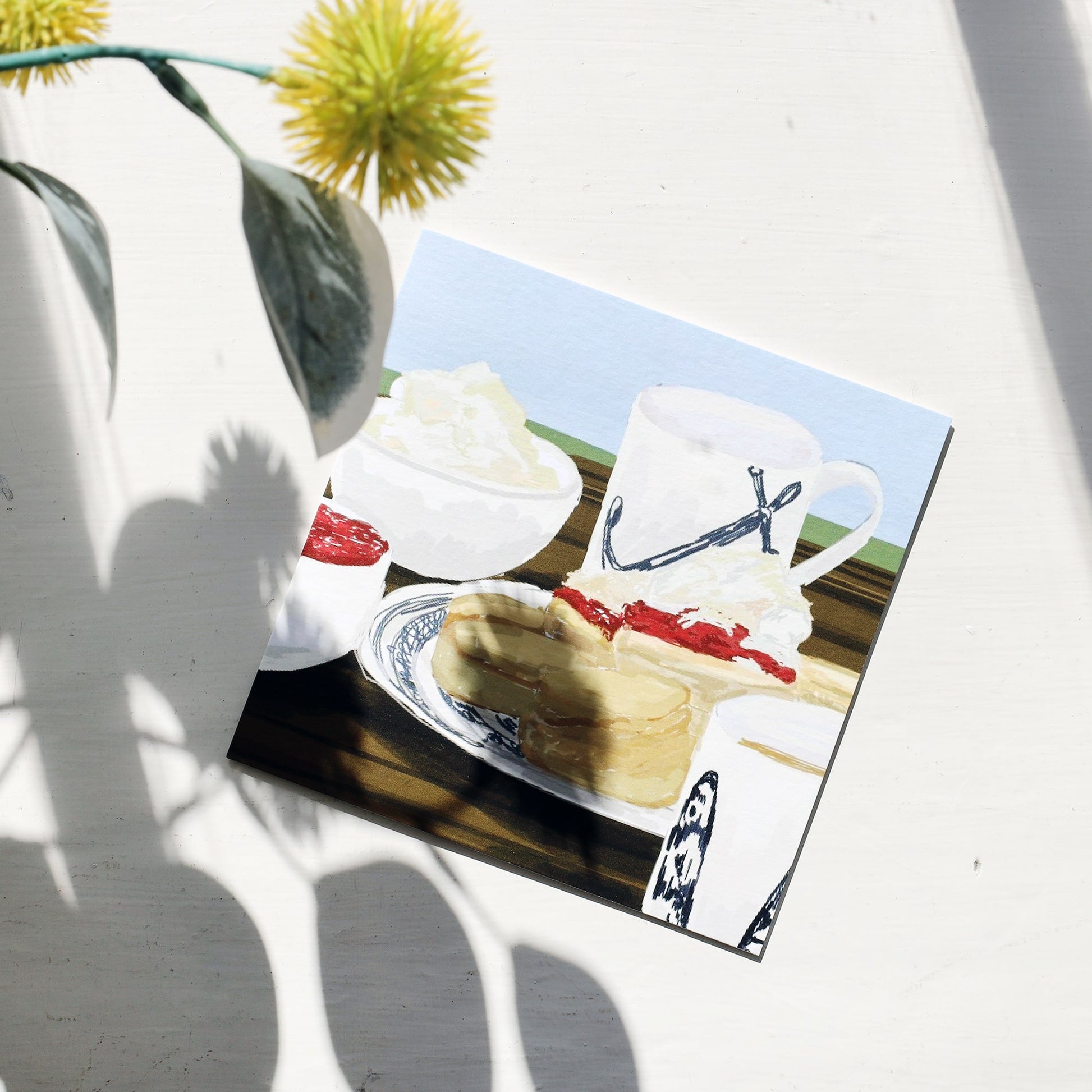 Greeting card featuring an illustrated cream tea scene with scones, jam, clotted cream, and a mug on a table, placed on a white surface with sunlight and shadows cast by a nearby plant.
