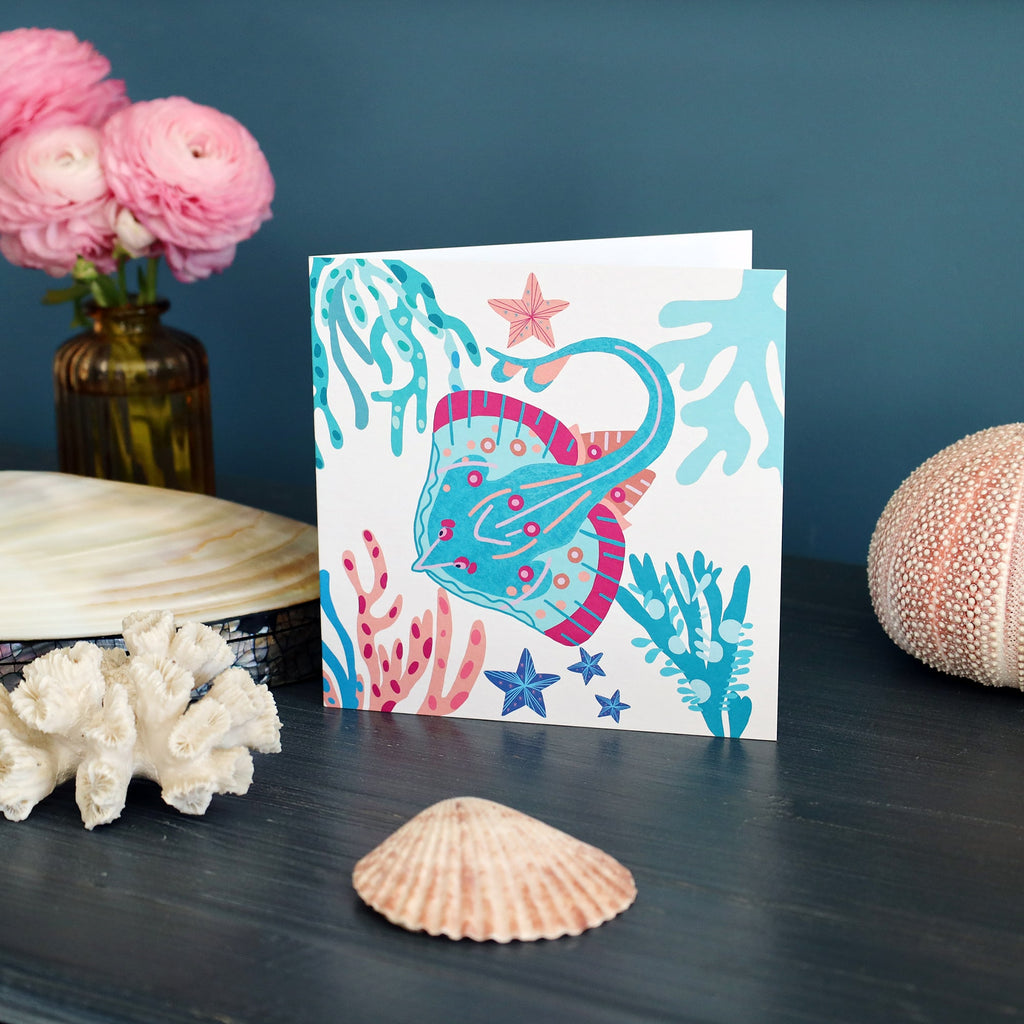 A brightly illustrated greeting card featuring a colorful stingray surrounded by coral, seaweed, and starfish in shades of turquoise, pink, and blue. The card is displayed upright on a dark wooden surface, accompanied by seashells, coral pieces, and pink ranunculus flowers in a glass vase, with a teal wall in the background.
