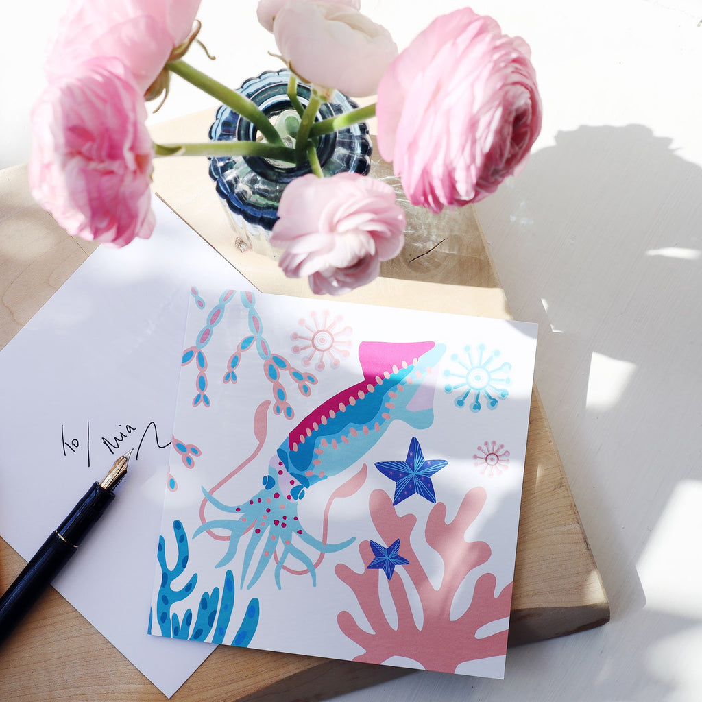 A vibrant square greeting card featuring a colorful squid, coral, starfish, and abstract sea shapes in shades of blue, pink, and purple. The card is placed on a light wooden surface next to a handwritten envelope, a black fountain pen, and a vase with pink ranunculus flowers. Sunlight casts soft shadows across the scene.