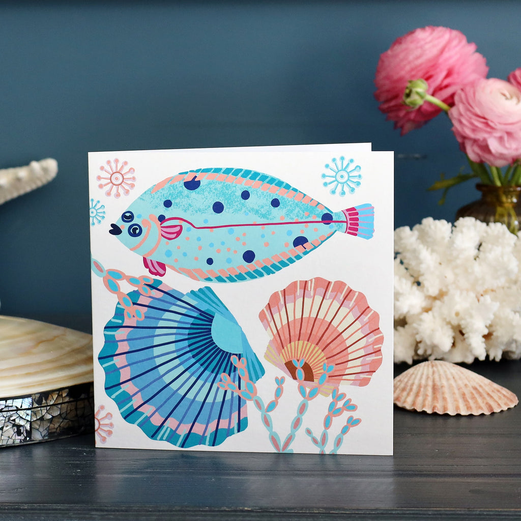 A bright and cheerful sea-themed greeting card is displayed upright against a dark blue background. The card features a playful illustration of a spotted flatfish, two scallop shells, and coral in shades of turquoise, pink, and orange. Surrounding the card are decorative coastal elements including a pink ranunculus bouquet in a glass vase, white coral, and a scallop shell, enhancing the seaside aesthetic.