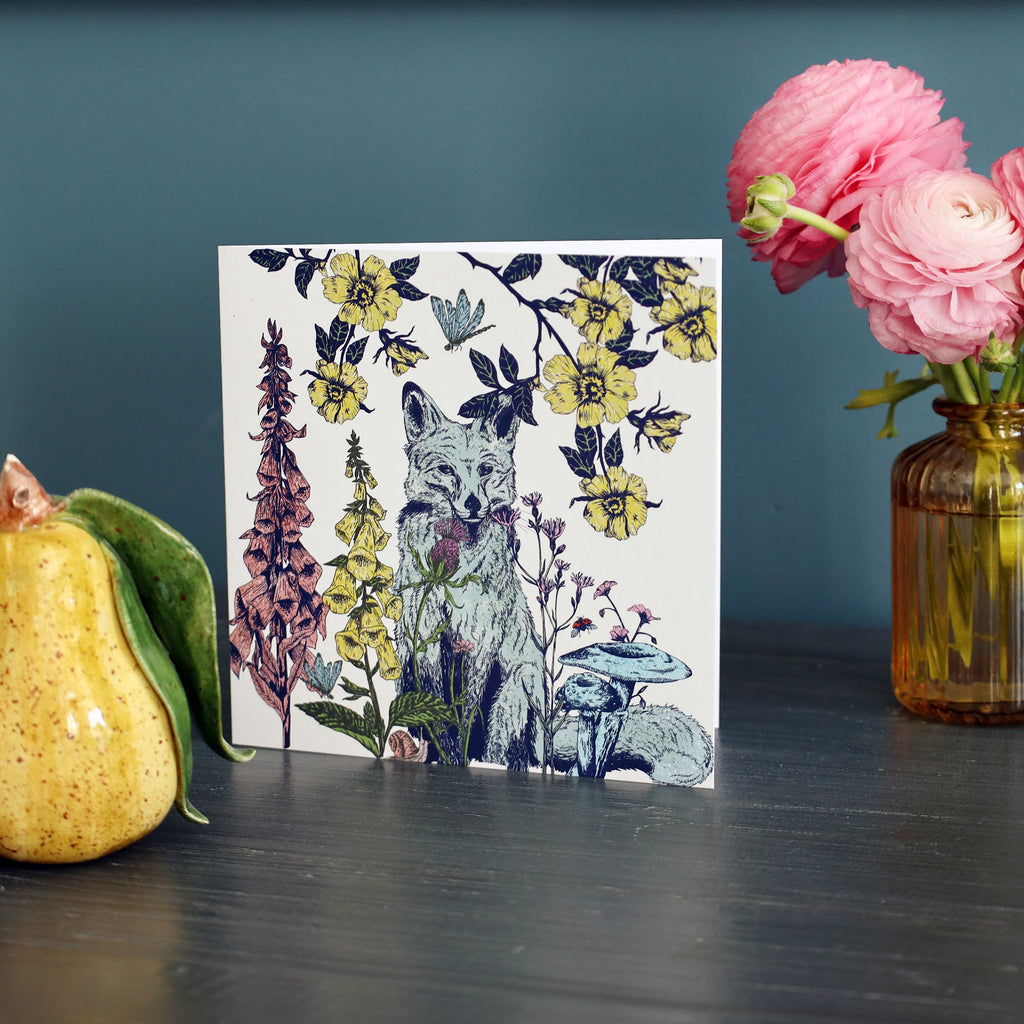A square greeting card featuring an illustrated fox surrounded by yellow flowers, wild plants, and mushrooms is displayed upright on a dark wooden surface. To the left is a ceramic pear ornament, and to the right is a vase of pink ranunculus flowers, set against a teal blue background.