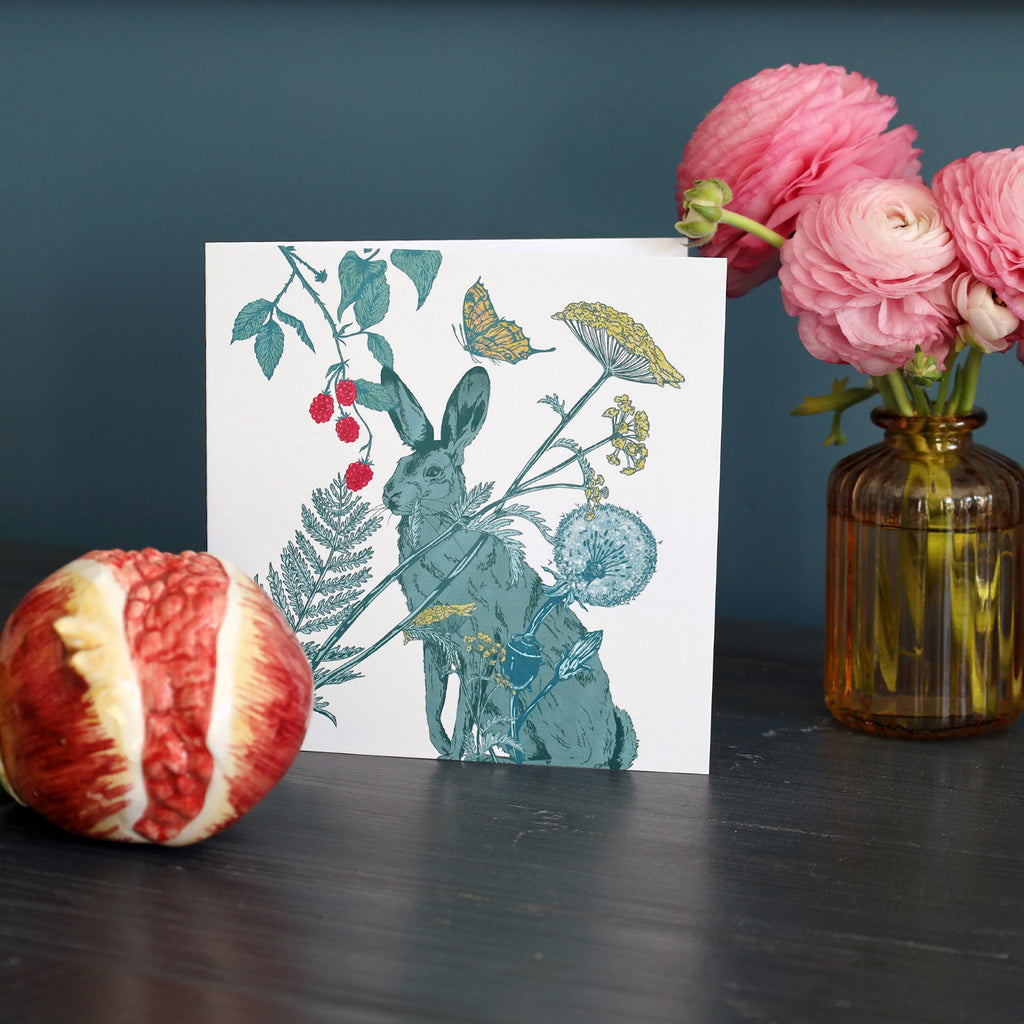 Greeting card featuring a detailed illustration of a hare surrounded by wildflowers, berries, ferns, and a butterfly. The card is set on a dark wooden surface, with a red ceramic pomegranate ornament on the left and a glass vase of pink ranunculus flowers on the right, against a muted teal background.
