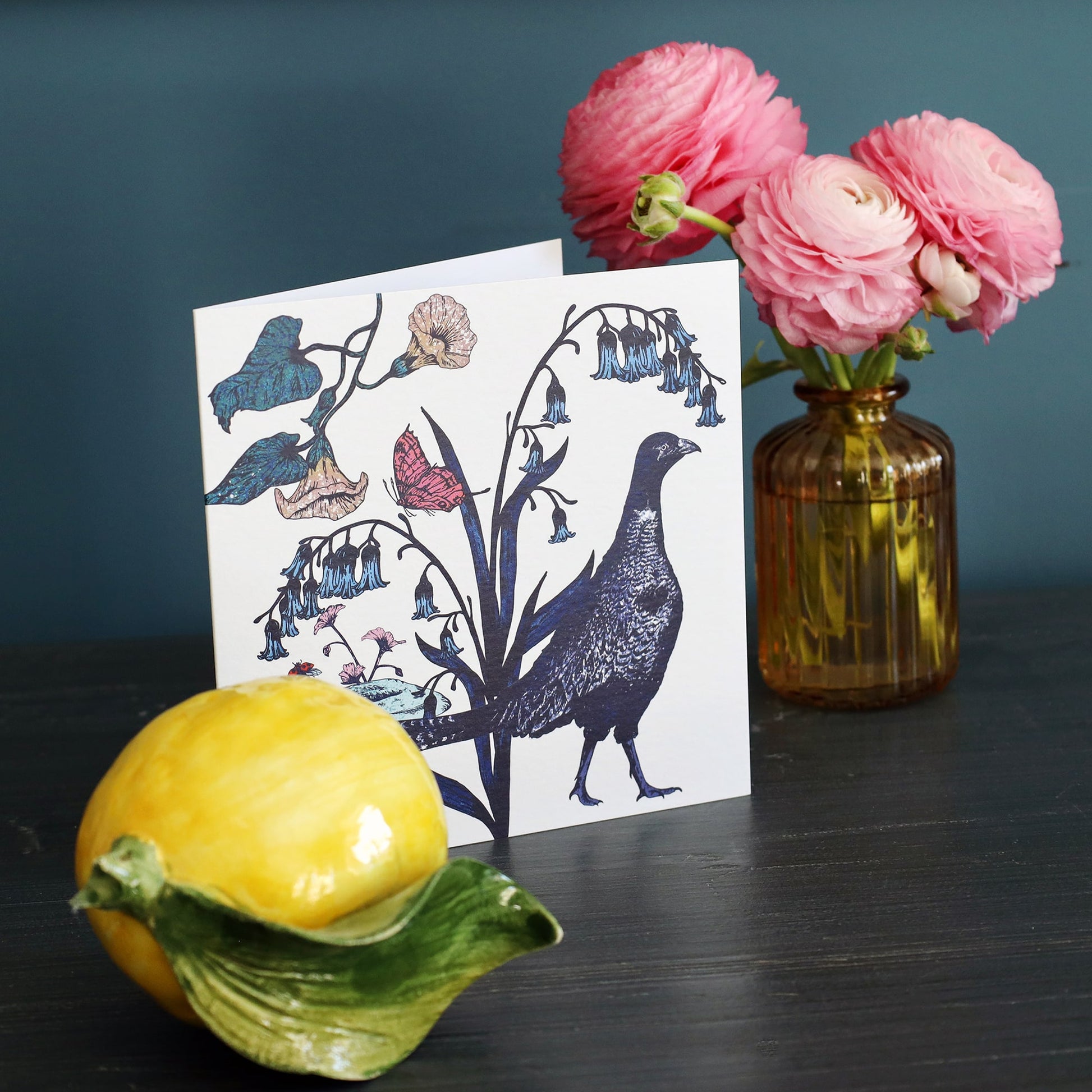 Illustrated pheasant greeting card standing on a dark wooden table next to a ceramic lemon and a glass vase filled with pink ranunculus flowers, set against a teal background.