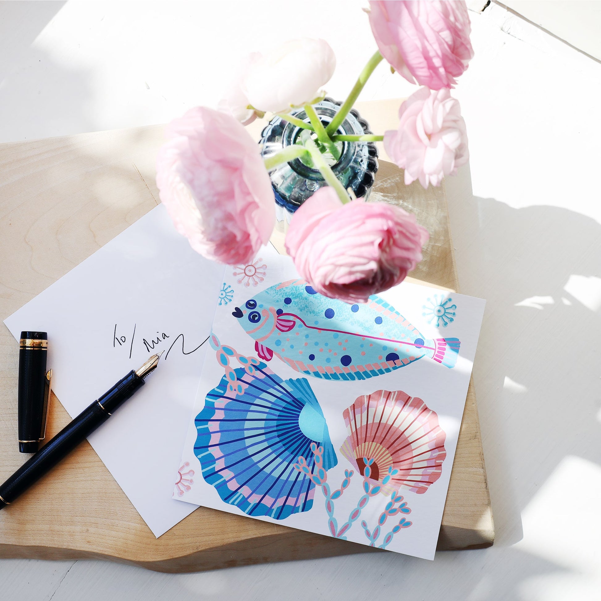 A colourful sea-themed greeting card featuring a spotted flatfish, scallop shells, and seaweed illustrations lies on a light wooden board. A fountain pen and an envelope addressed "To Mia" are beside it, with a vase of pink ranunculus flowers casting soft shadows over the scene.




A colourful sea-themed greeting card featuring a spotted flatfish, scallop shells, and seaweed illustrations lies on a light wooden board. A fountain pen and an envelope addressed "To Mia" are beside it, with a vase of pink r


