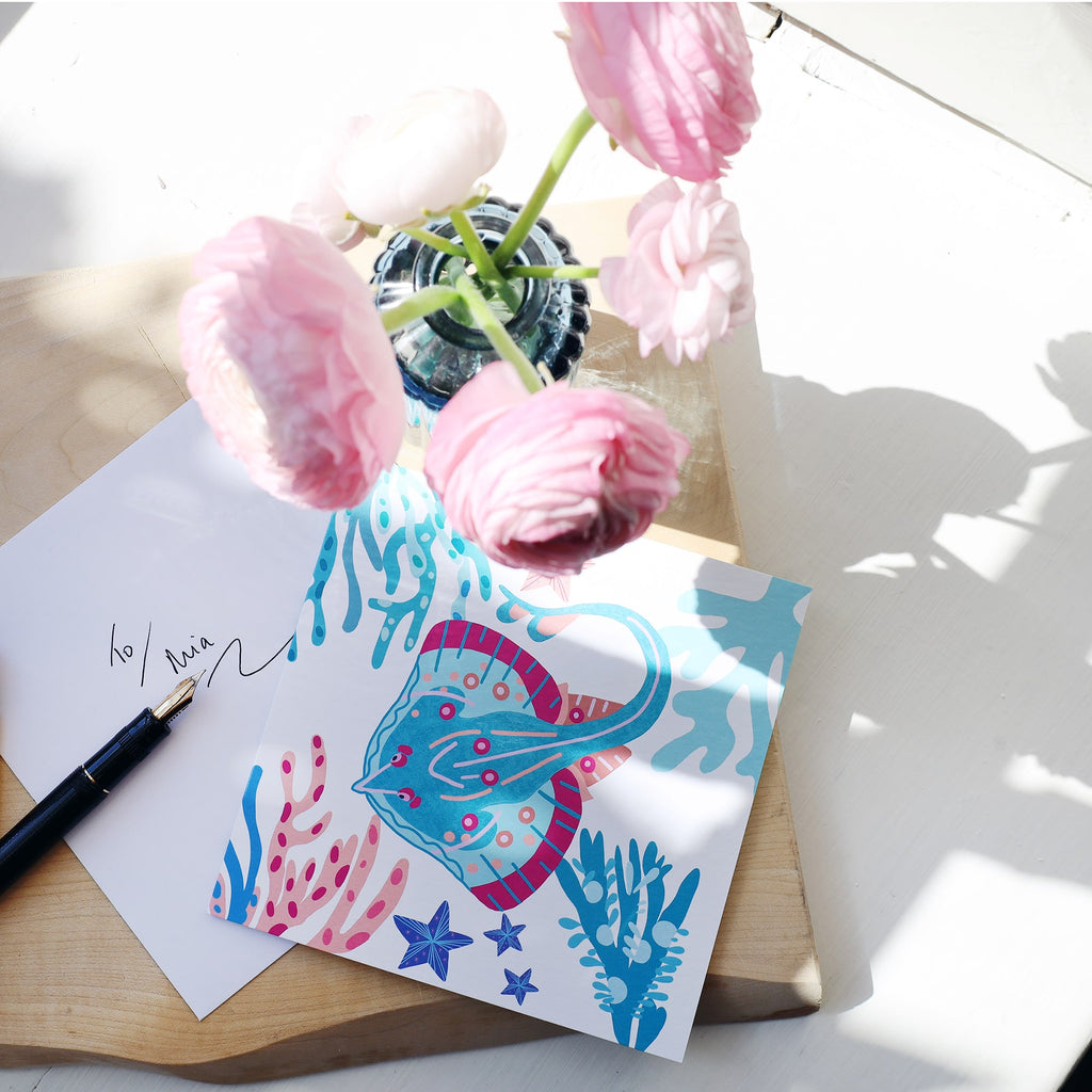 A vibrant greeting card featuring a colorful stingray surrounded by underwater plants and starfish, in shades of blue, teal, and pink. The card is styled on a wooden board with a handwritten envelope, a black fountain pen, and a vase of pink ranunculus flowers, all bathed in soft natural light.