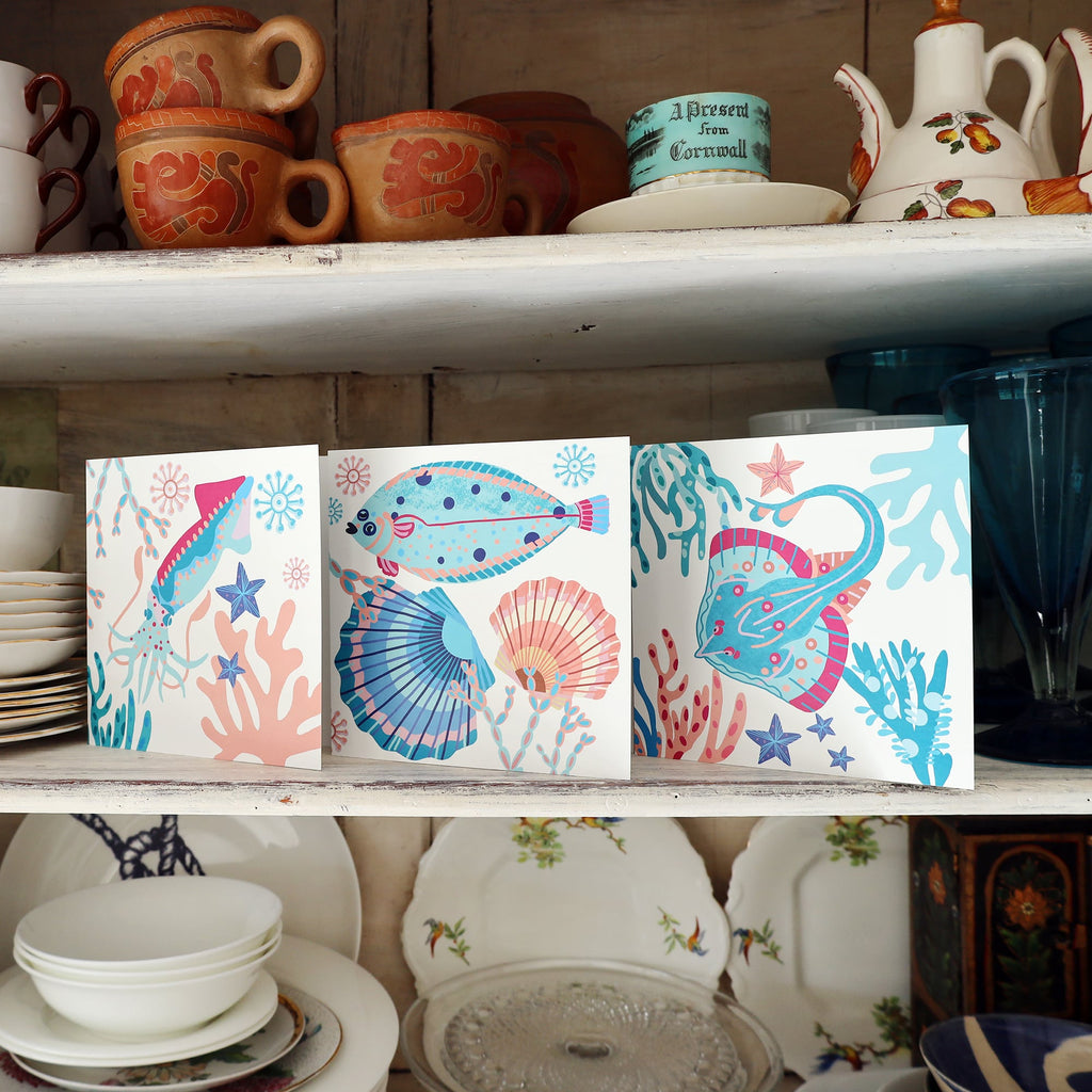 Three colorful, sea-themed greeting cards are displayed on a white wooden shelf in a vintage kitchen setting. Each card features vibrant illustrations of marine life including a squid, flatfish, stingray, seashells, and coral in shades of pink, blue, and turquoise. Surrounding the cards are stacks of crockery, vintage mugs, teapots, and glasses, adding a nostalgic and homely feel.