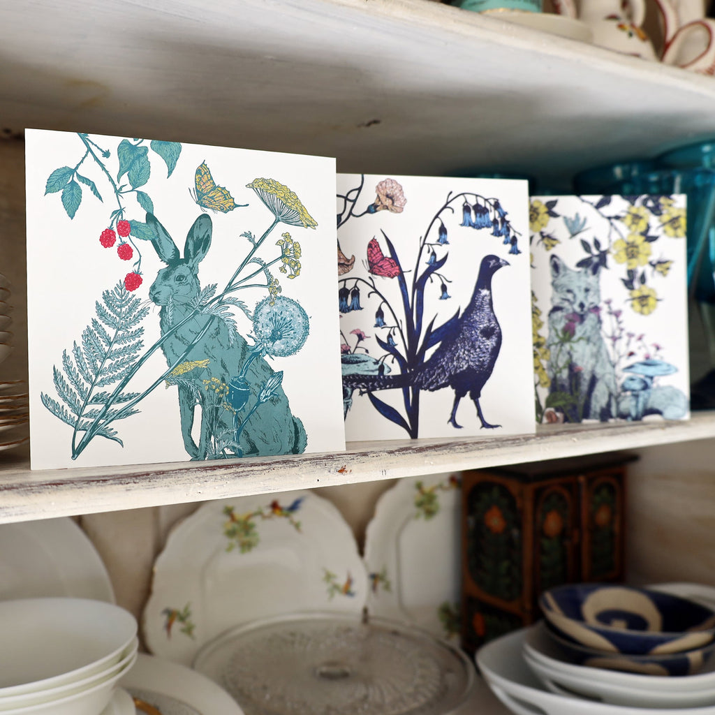 A trio of illustrated greeting cards displayed on a vintage white wooden shelf. The first card features a hare surrounded by wildflowers and berries, the second card depicts a pheasant standing among bluebells and butterflies, and the third card shows a fox nestled within foliage and mushrooms. Below the shelf are delicate floral-patterned plates and bowls, creating a charming and rustic backdrop.