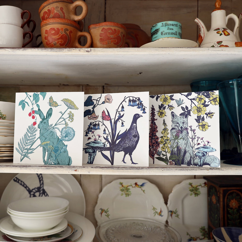 A trio of illustrated greeting cards displayed on a wooden shelf. The first card features a hare surrounded by wildflowers, berries, and butterflies. The middle card shows a pheasant standing among bluebells and mushrooms with butterflies overhead. The third card depicts a fox surrounded by foxgloves, yellow flowers, mushrooms, and a dragonfly. Vintage crockery and glassware are arranged on shelves around the cards.