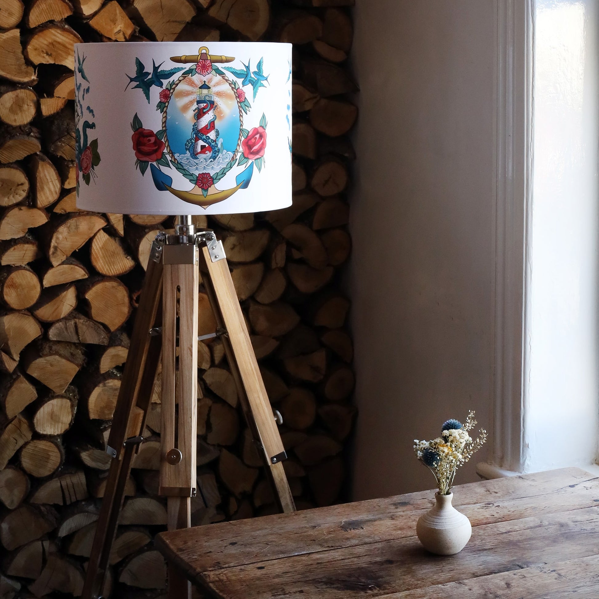 Lampshade on wooden floor tripod with lighthouse and kraken design from our Sailor's Story collection. The tripod is standing in front of a stack of cut logs and there is a small wooden table with a vase and dried flowers.
